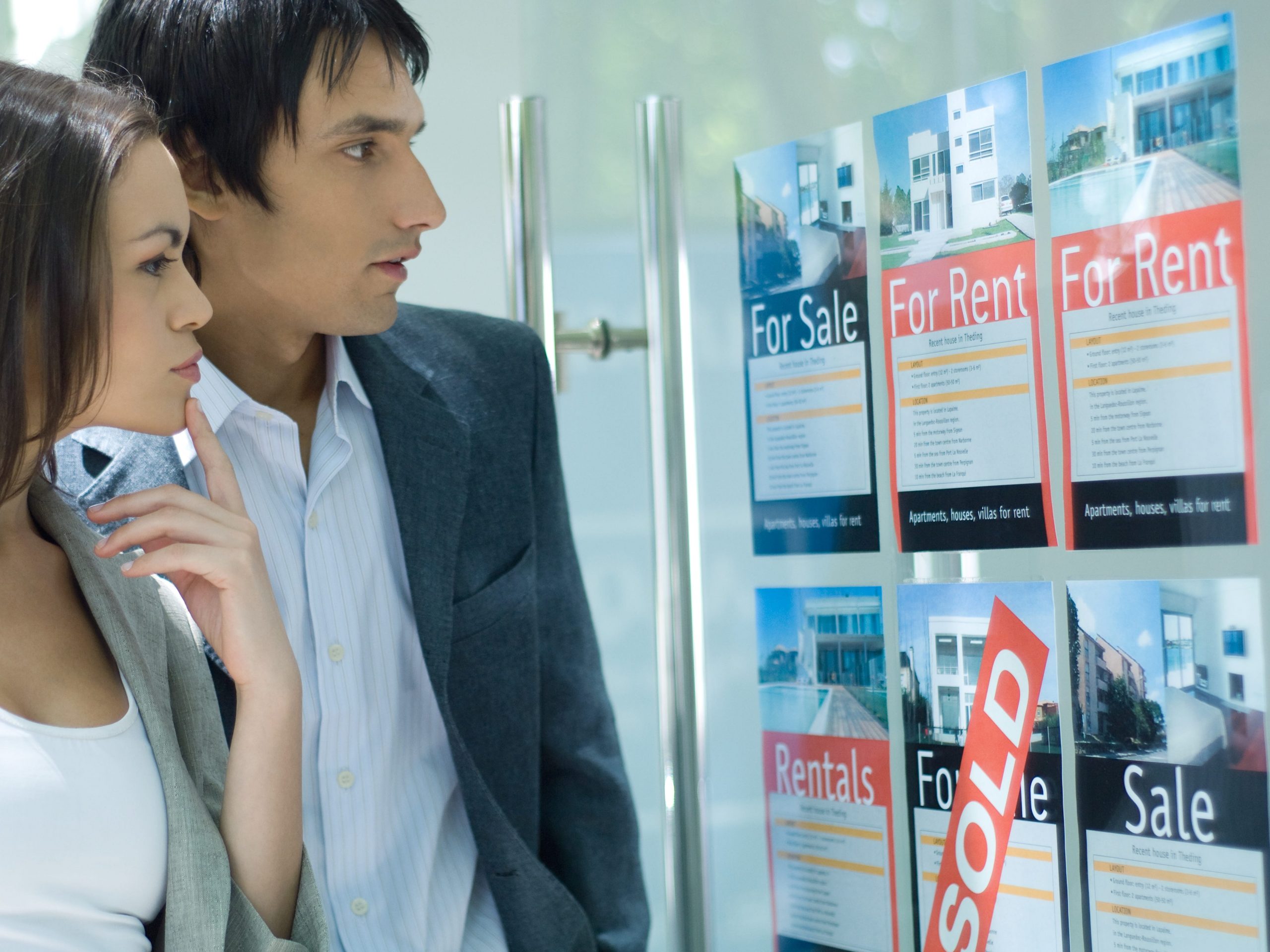 Couple standing next to window of real estate agency, looking at for rent and for sale signs.