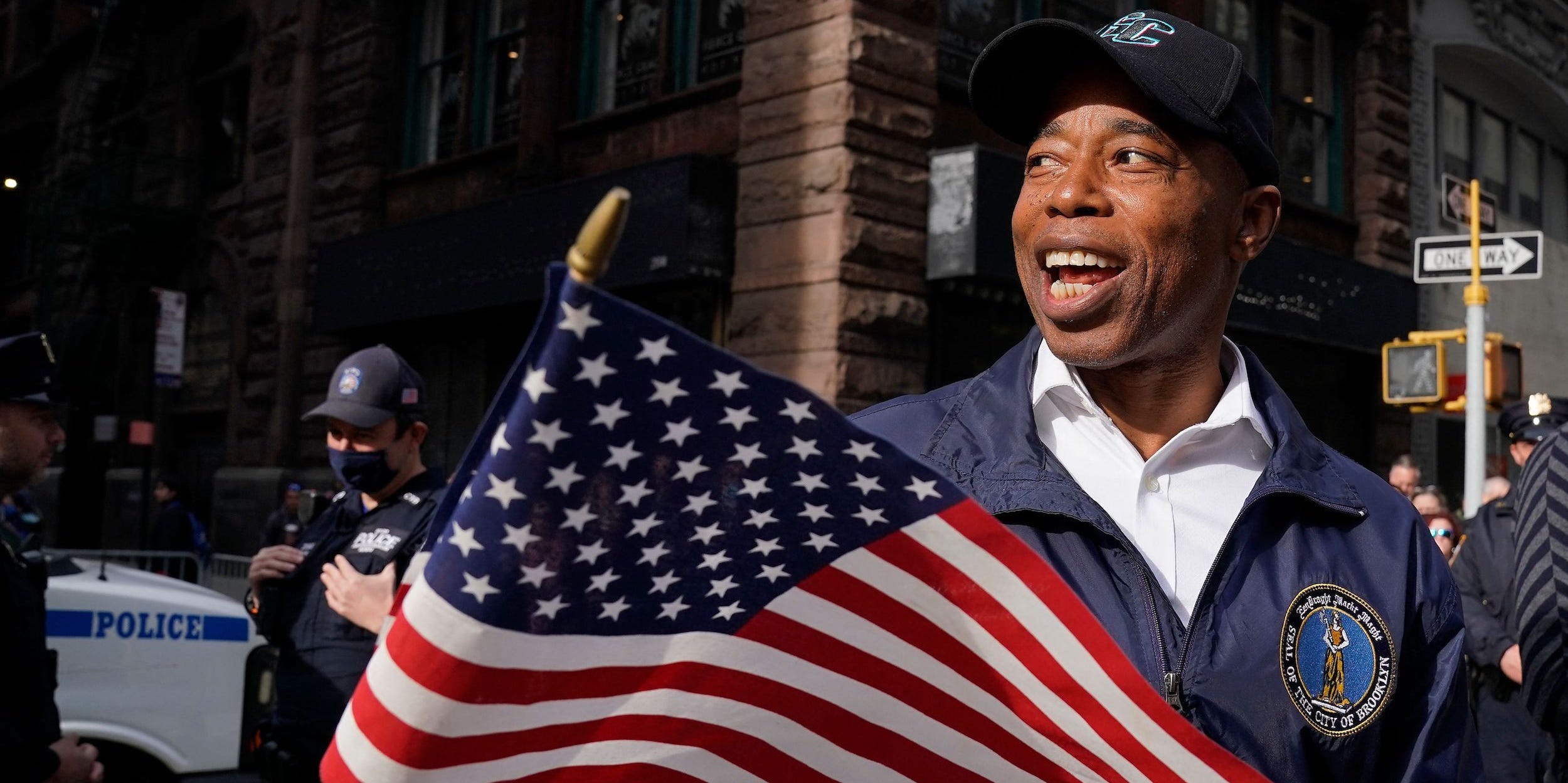 Eric Adams waves a slightly larger than miniature but not quite as big as a full-sized American flag while wearing a baseball cap and a Brooklyn borough windbreaker.