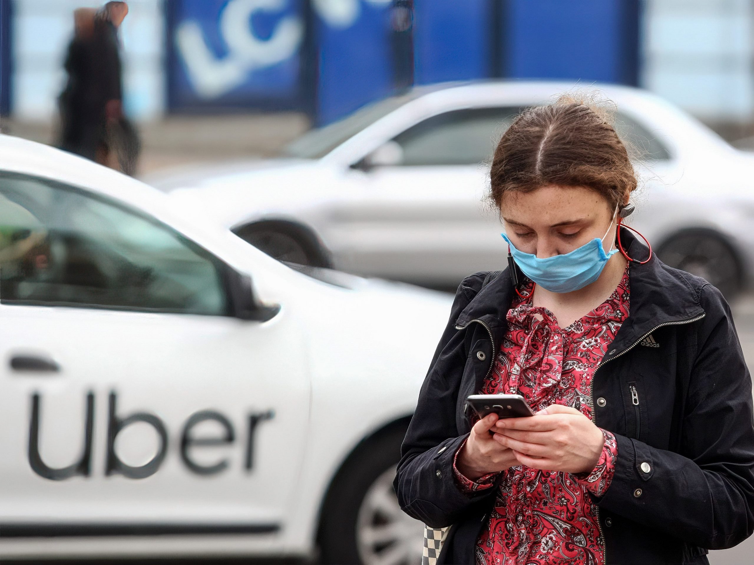 A woman with a mask on looks at her phone outside of an Uber car.