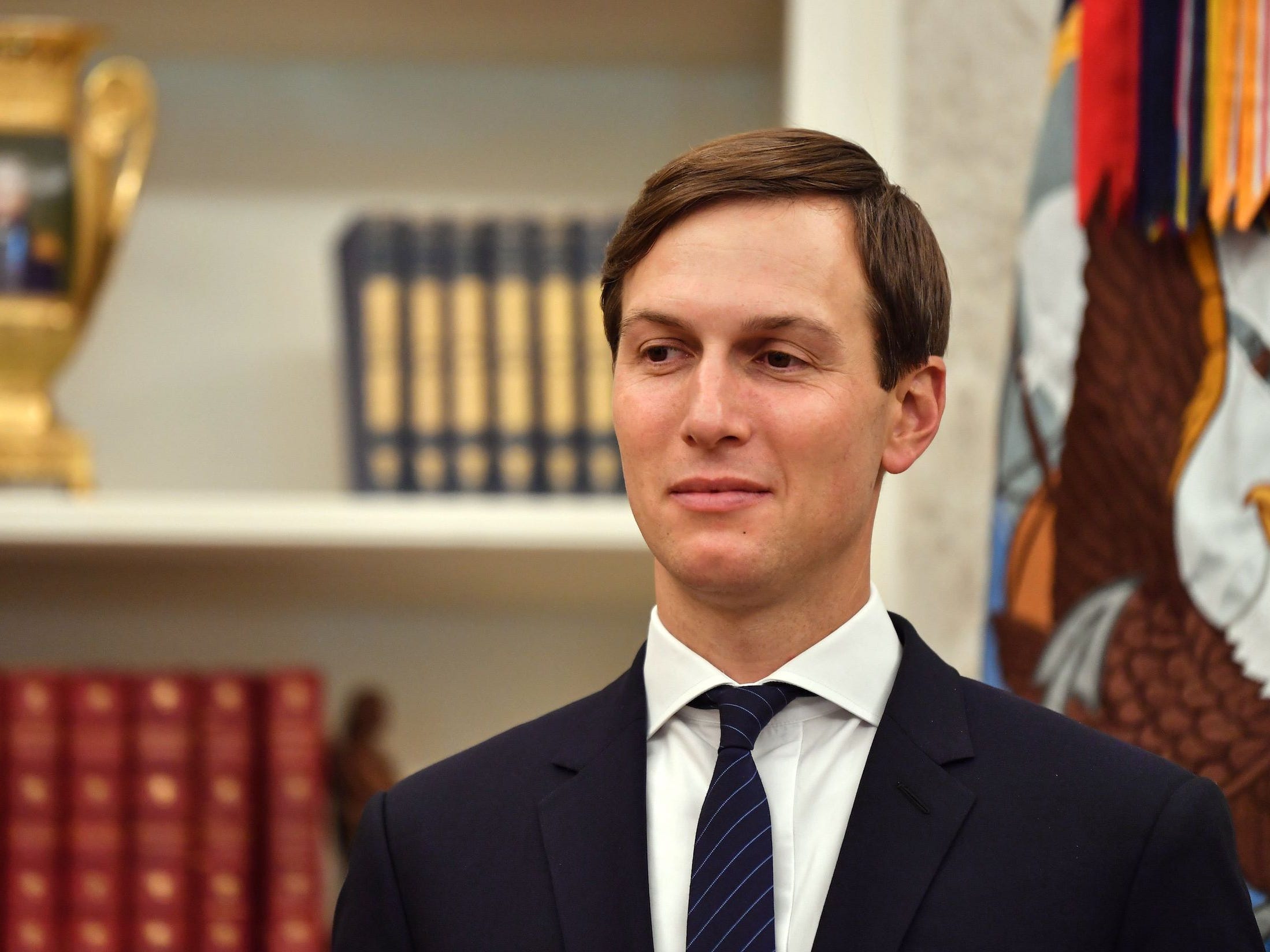 Jared Kushner stands in the oval office in front of a bookshelf with a golden chalice and two collections.