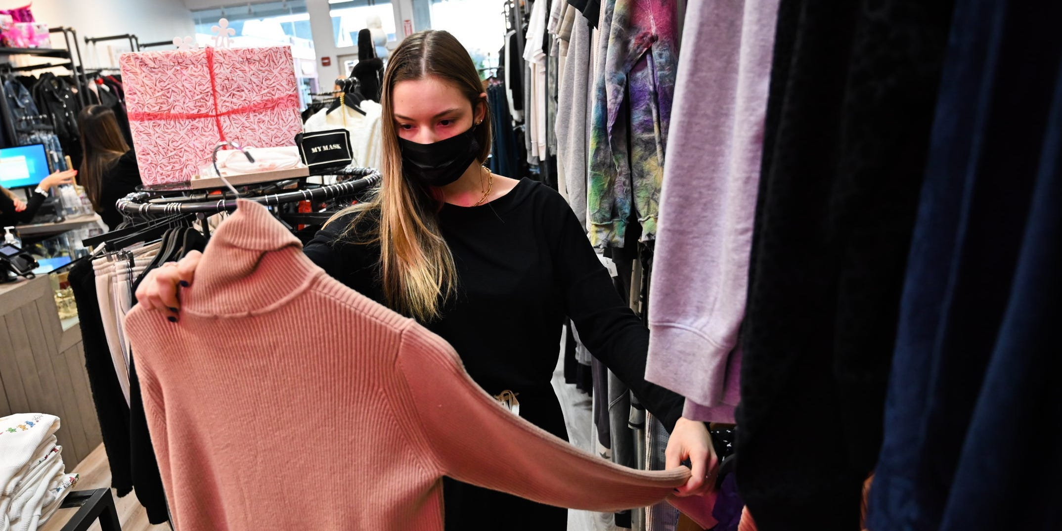 Woman wearing face mask holds up pink turtleneck sweater while clothes shopping