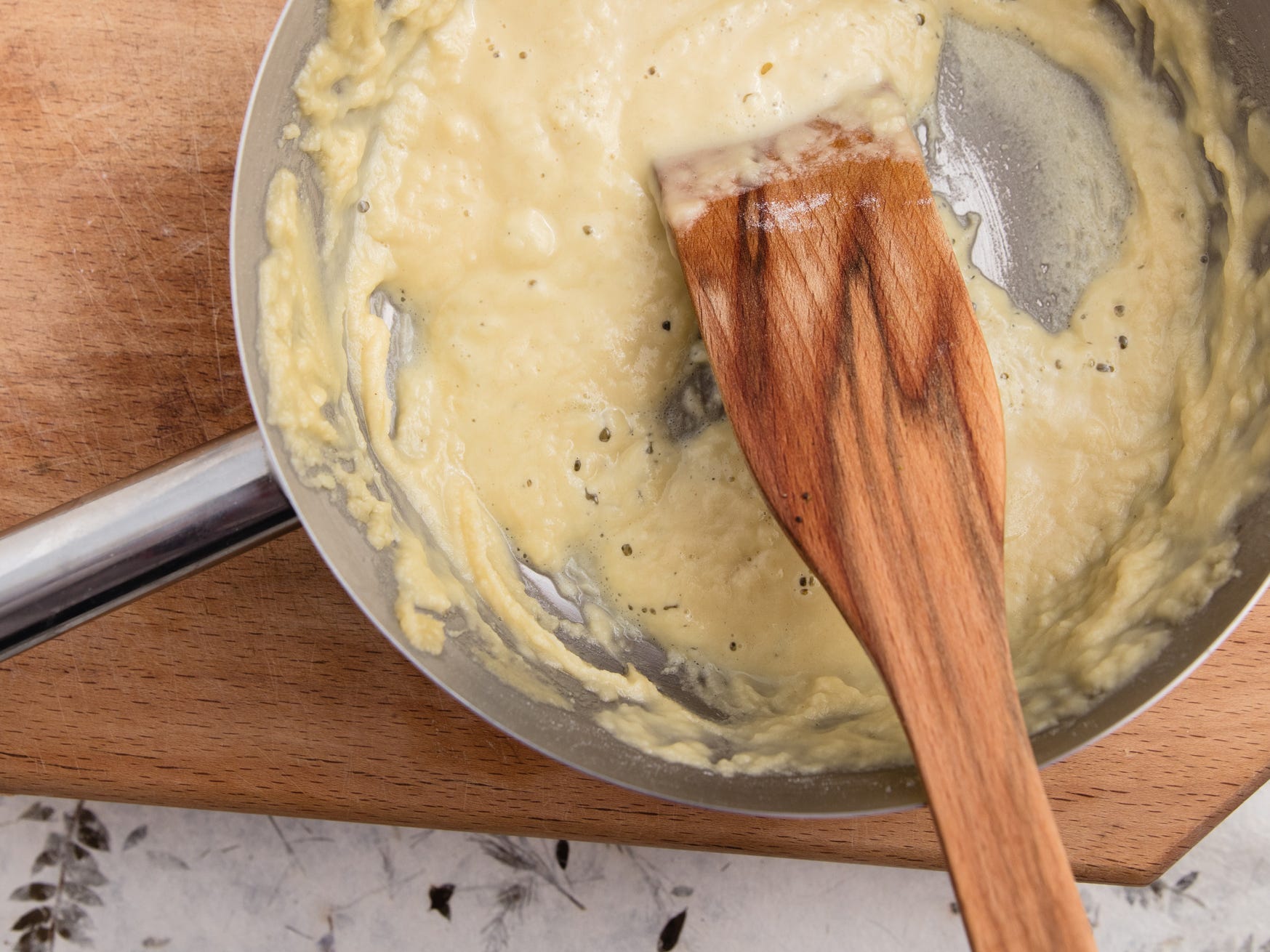 A pan of white roux sitting on a wooden cutting board being stirred with a wooden paddle