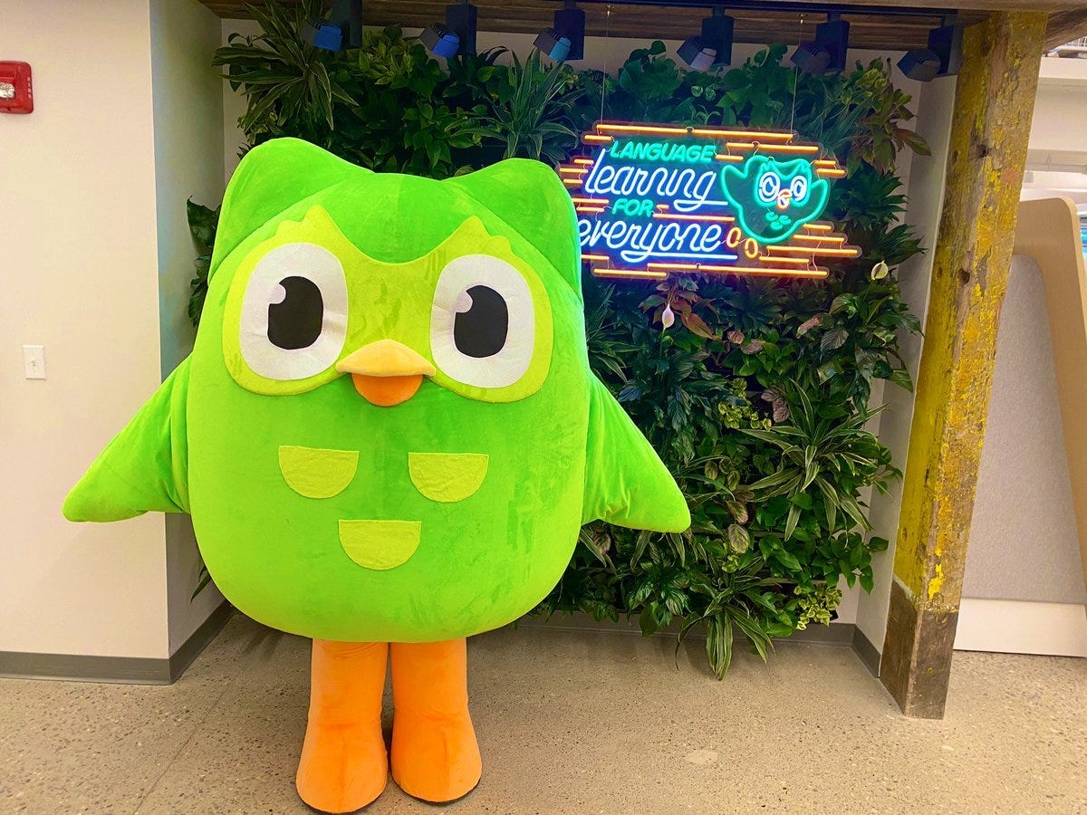 a person in costume as the duolingo bright green owl in front of a plant wall and a neon sign that says language learning for everyone