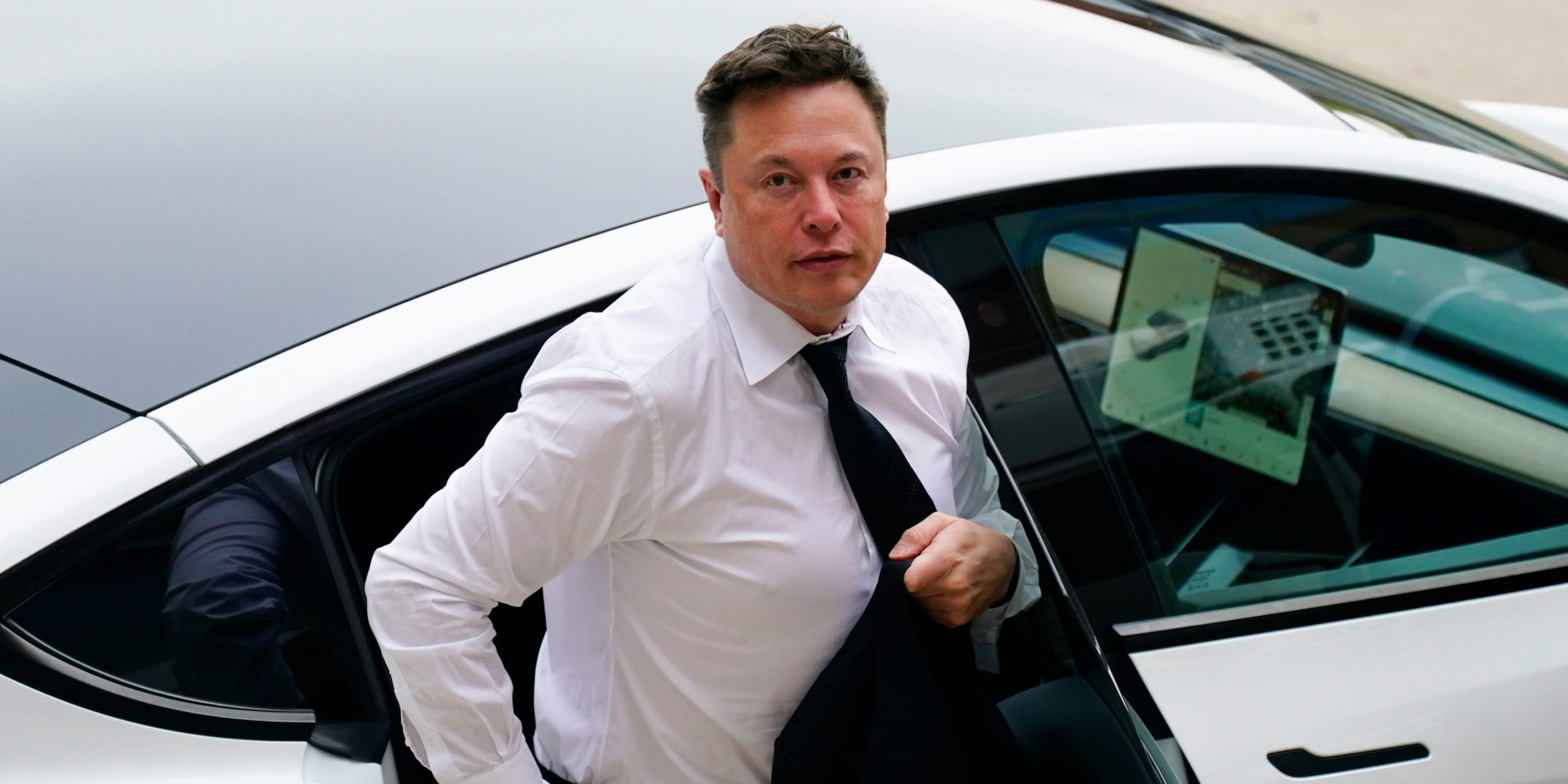 Tesla CEO Elon Musk stepping out of a silver Tesla wearing a white shirt and black tie on a sunny day