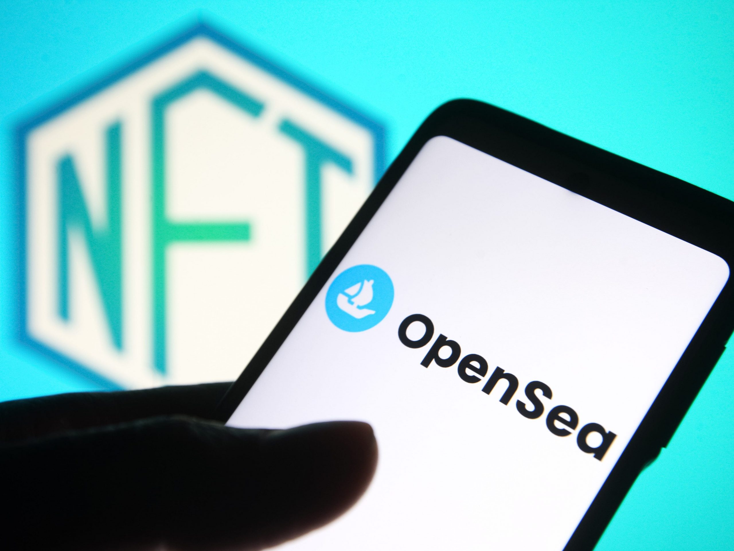 An OpenSea logo of an online marketplace for non-fungible tokens is seen on a smartphone screen and a NFT (Non-fungible token) sign in the background.