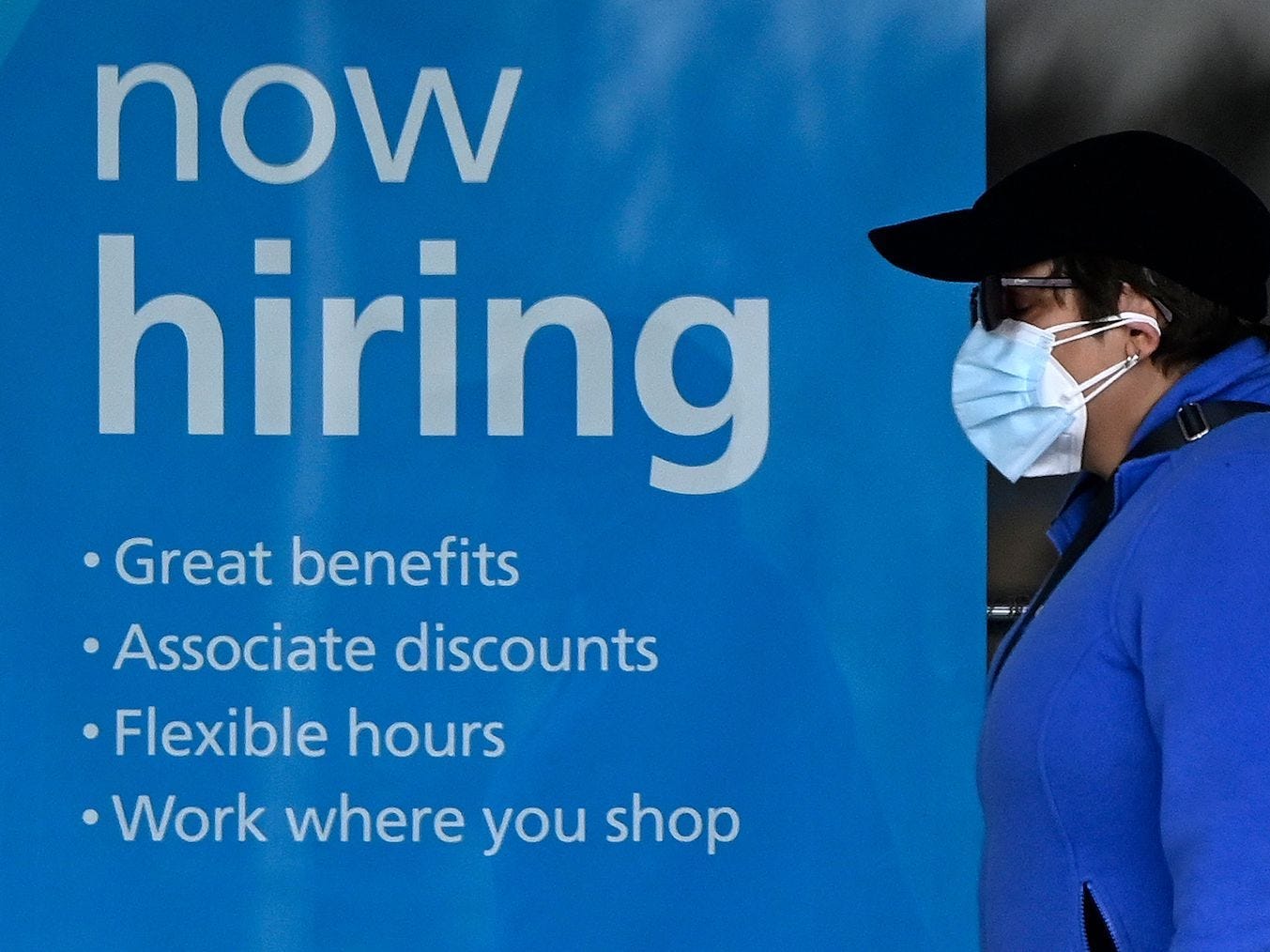 A woman wearing a face mask walks past a "Now Hiring" sign in front of a store.