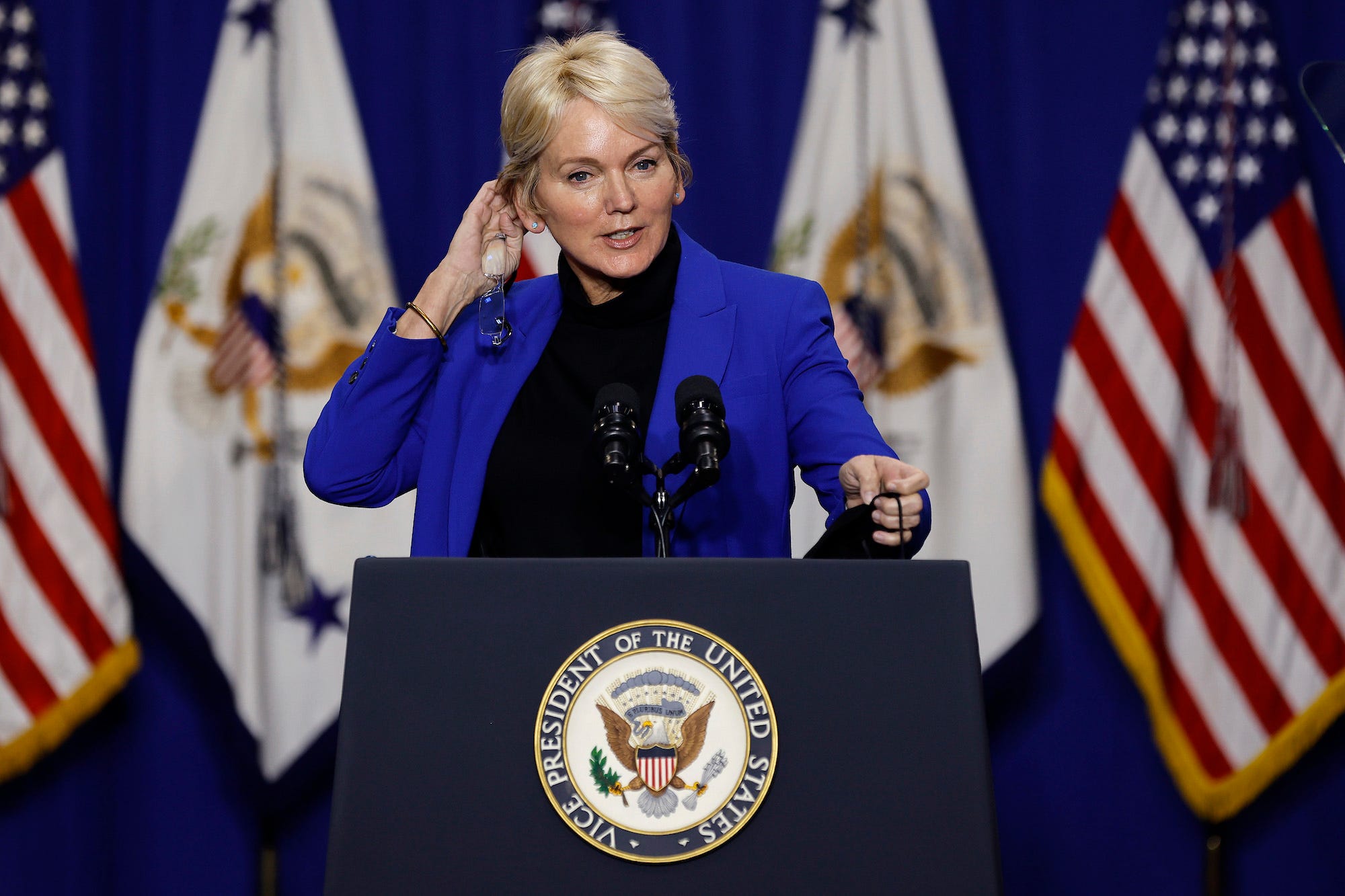 Energy Secretary Jennifer Granholm fixes her hair with right hand while standing against a backdrop of various flags at a Biden administration press conference.