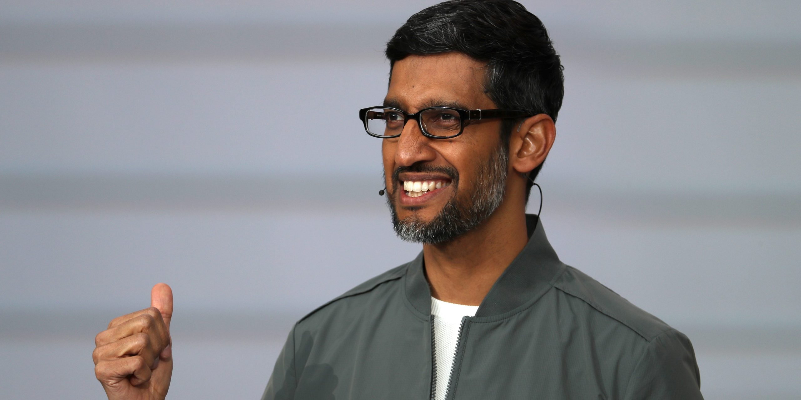 Sundar Pichai wears a grey jacket over a white t-shirt and smiles on stage.