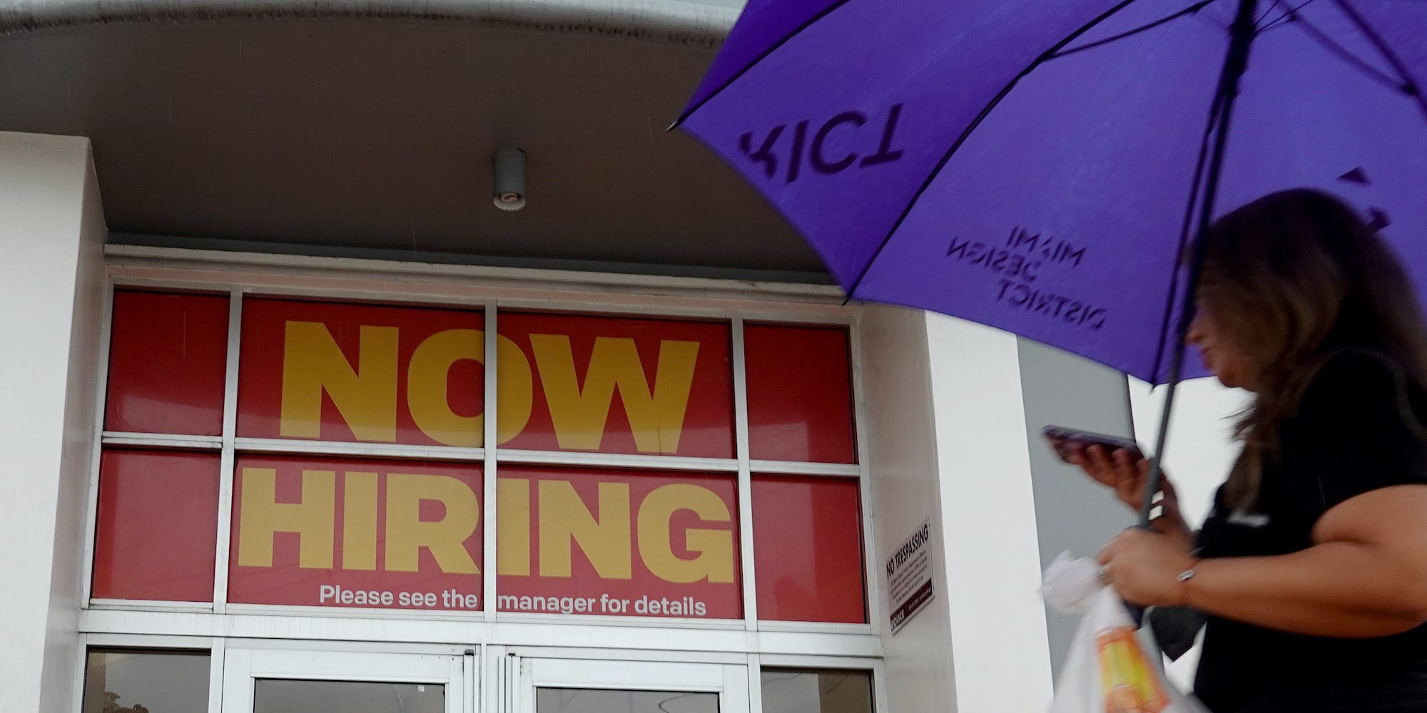 A person with an umbrella walks by a McDonald's restaurant that has a ''Now Hiring" sign above the entrance.