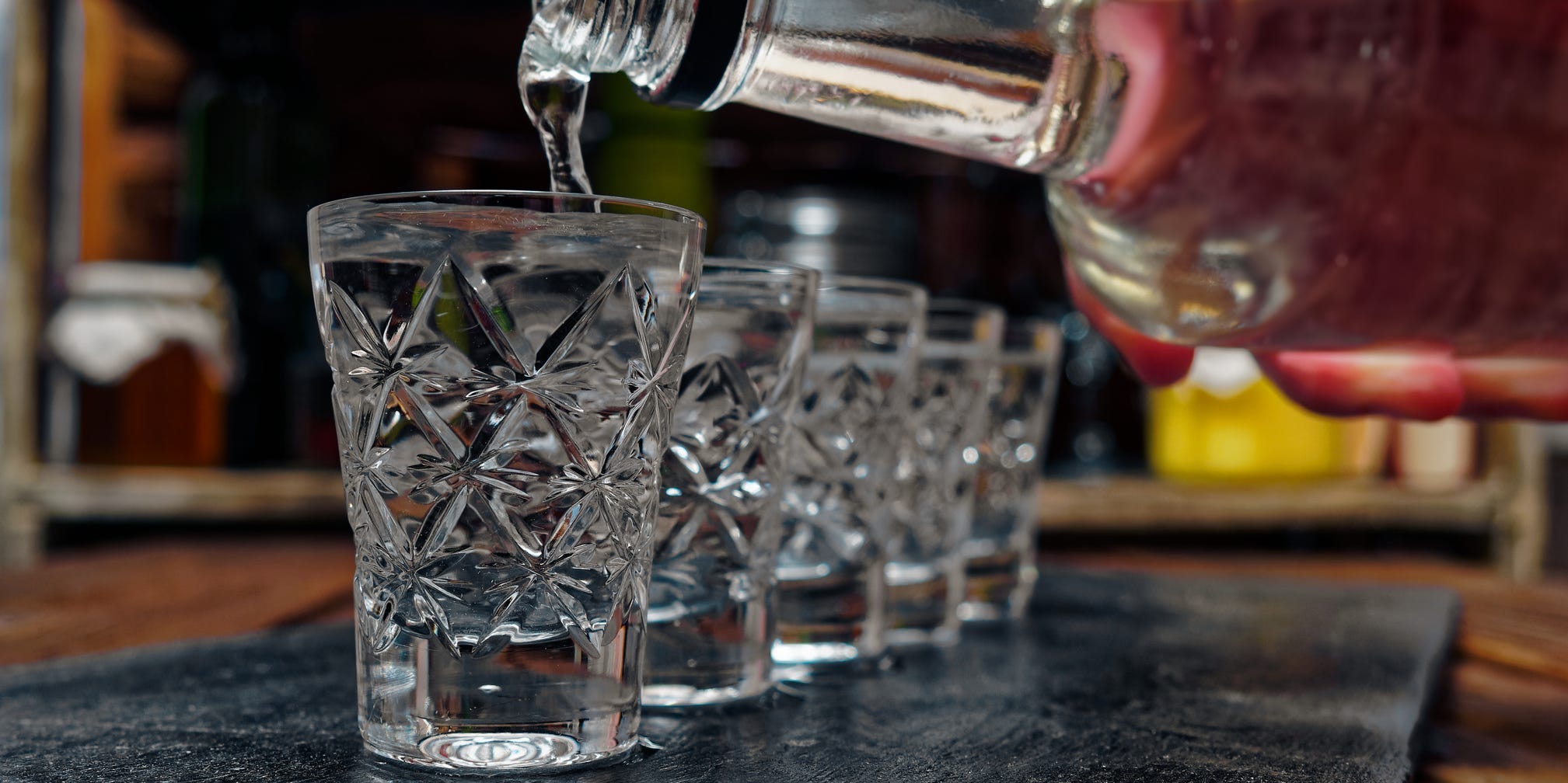 A bottle of vodka being poured into a line of shot glasses