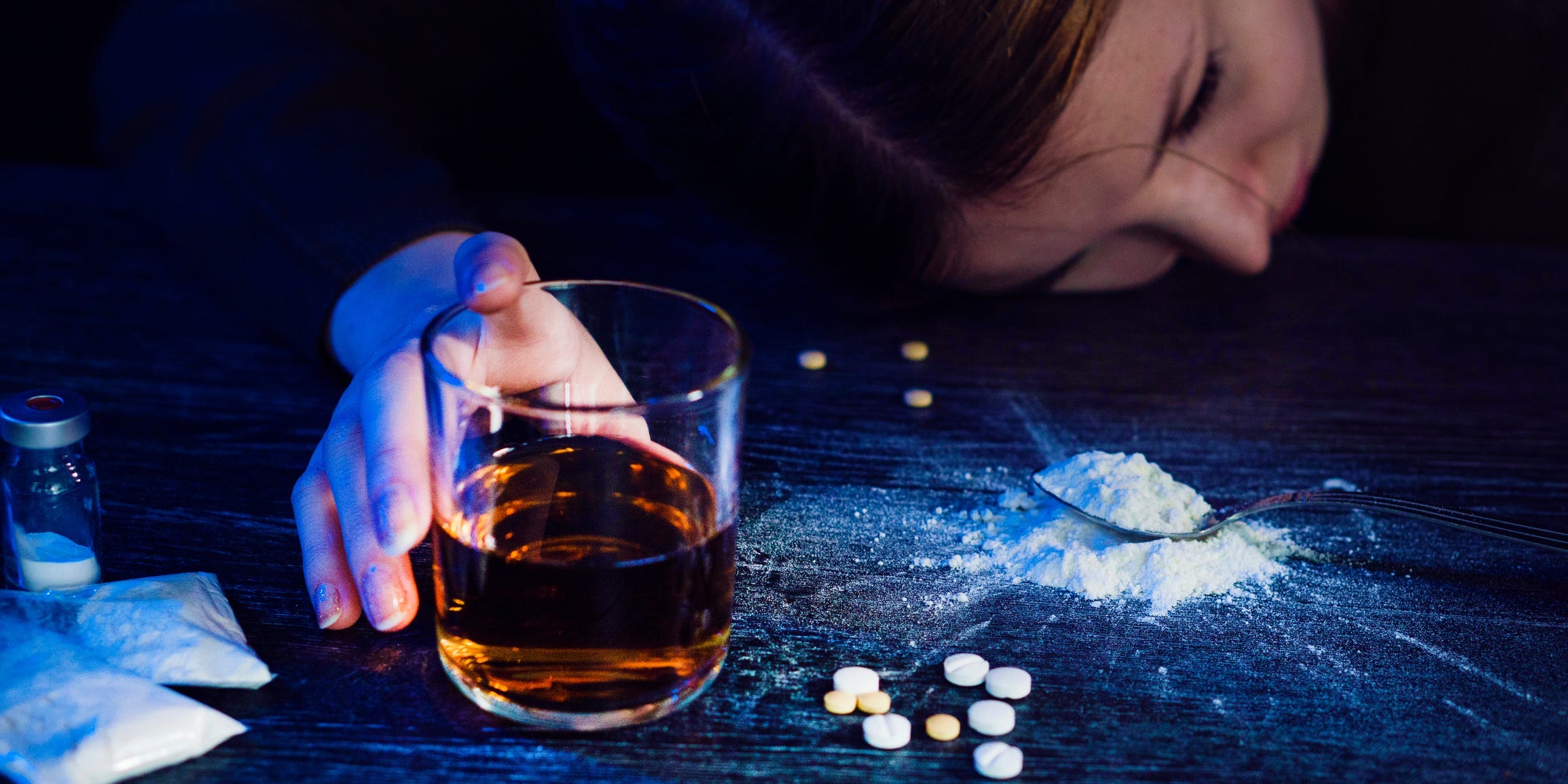 A person lies passed out on the floor with an unfinished glass of whiskey and some pills in front of them.