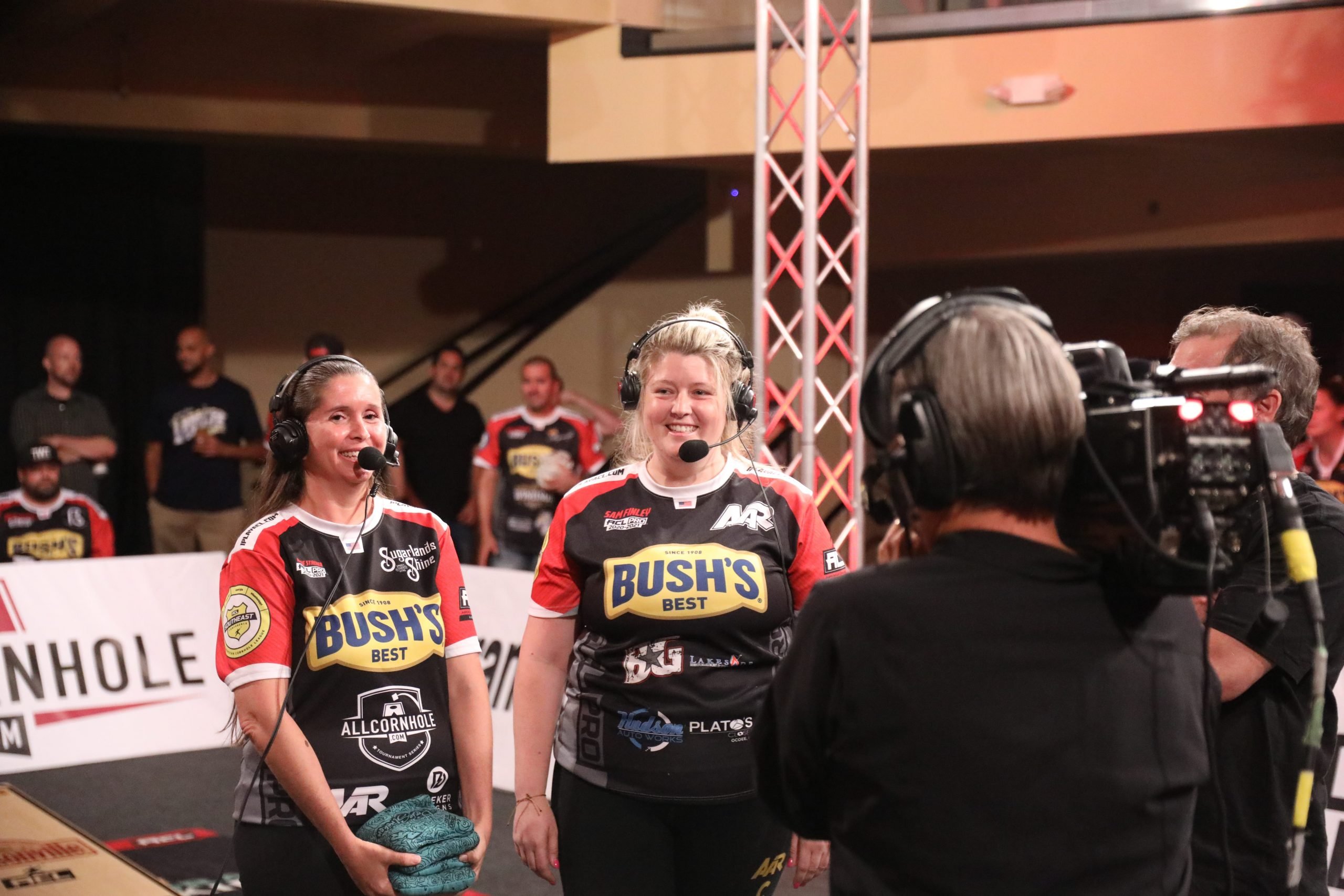 Two female COrnhole players wearing headsets stand in front of a camera