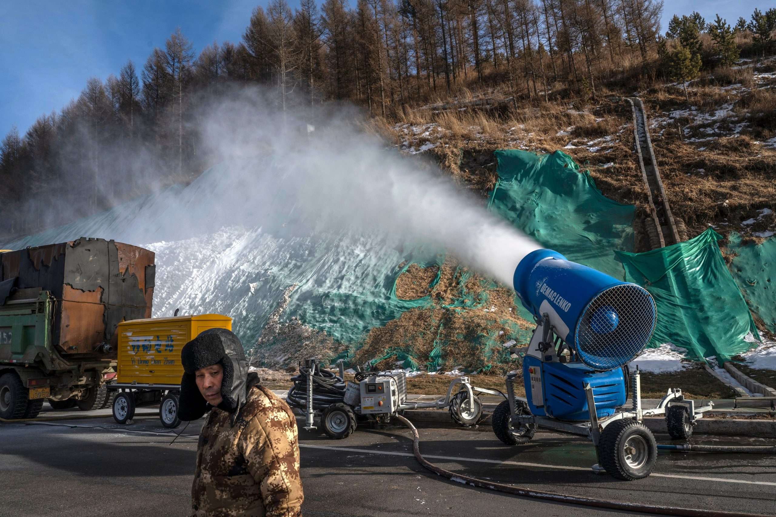 A worker stands next to a snow machine making artificial snow outside one of the athletes villages