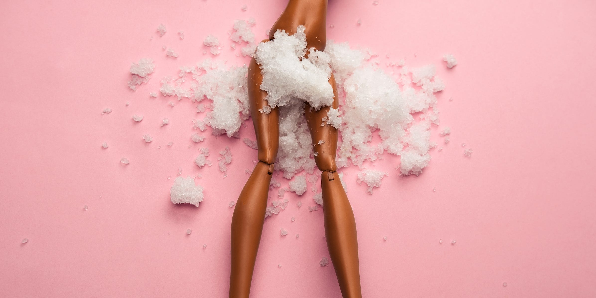 A doll on a pink background, seen from the waist down, has snow covering its hips.