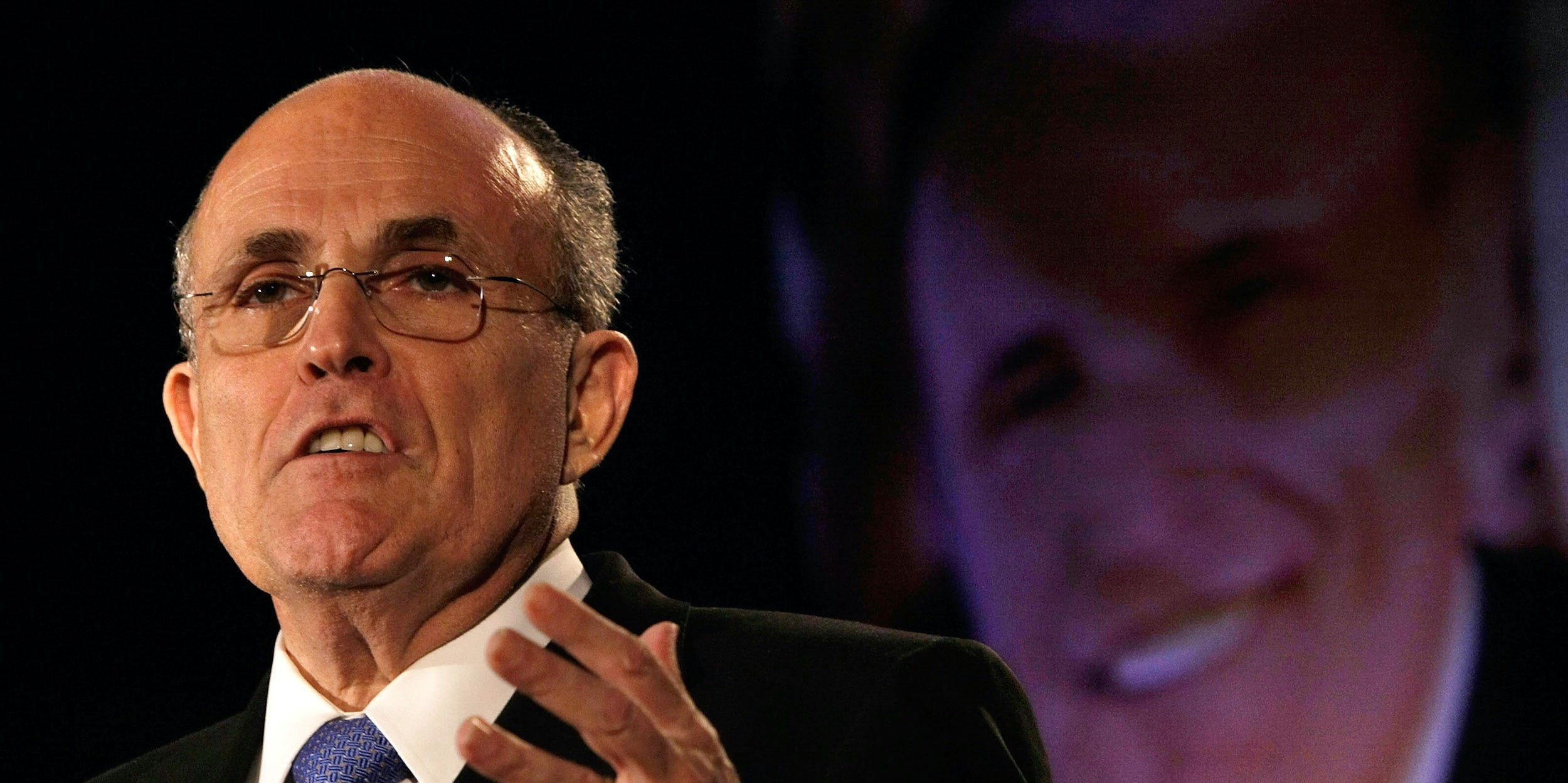 Rudy Giuliani speaks in front of a projected photo of his face.