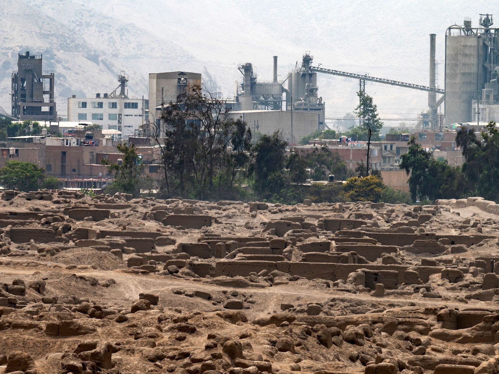 A view of Cajamarquilla, an archeological site outside of Lima, Peru, shows the excavated landscape in front of a backgorund of industrial buildings.