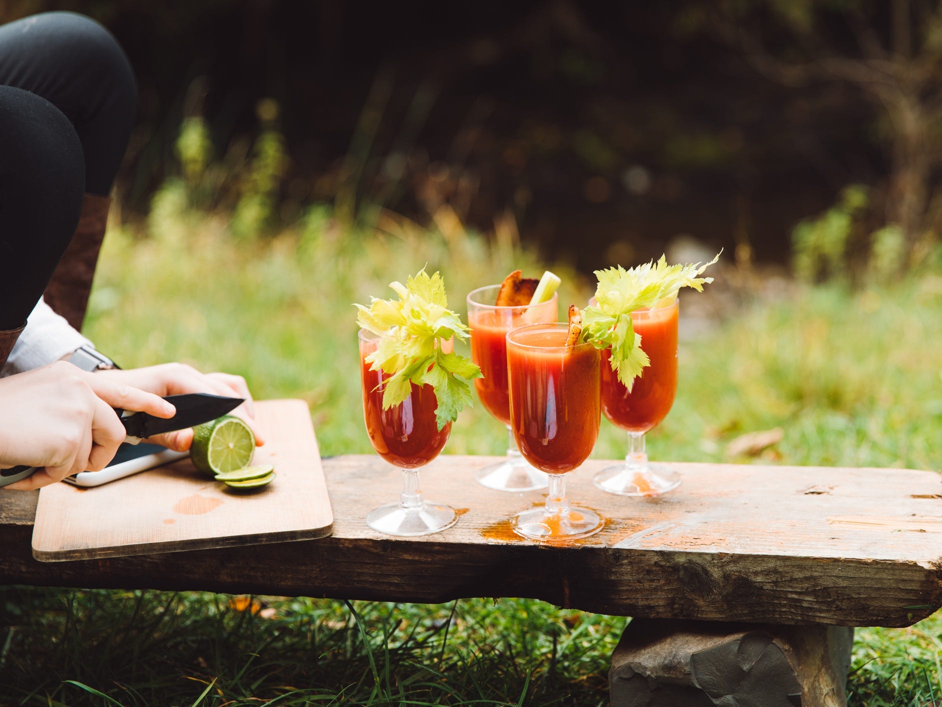Woman slicing a lime on a cutting board outside with four glasses of bloody marry cocktails