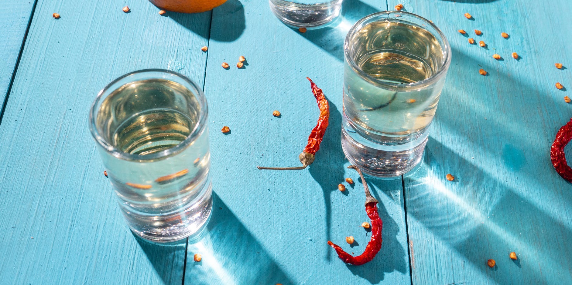 Mezcal shots with scattered orange segments and dried peppers on a bright blue wooden table