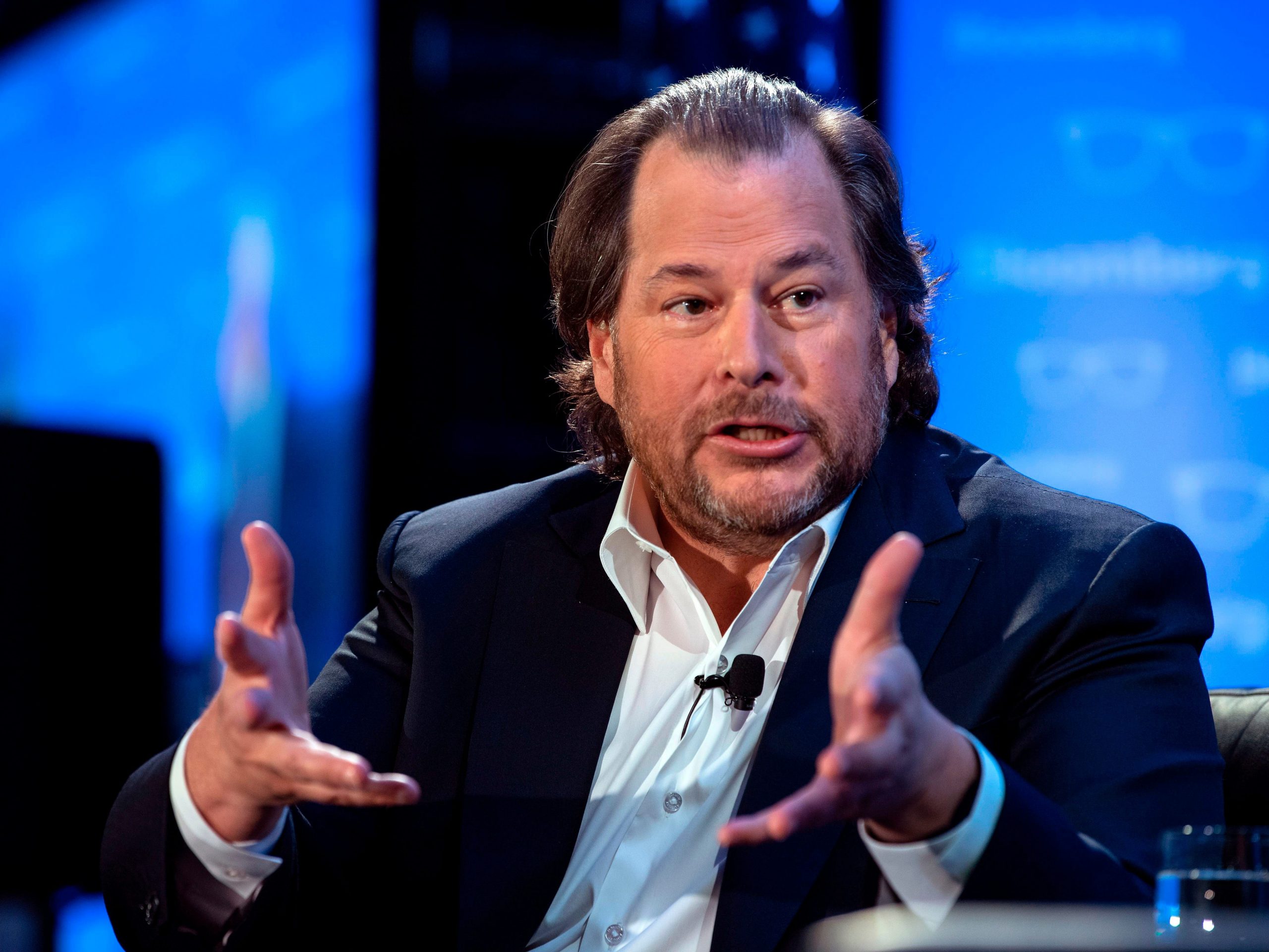 Salesforce founder Marc Benioff, wearing a sports coat jacket and in front of a blue background on a stage, extends his hands forward during a talk.