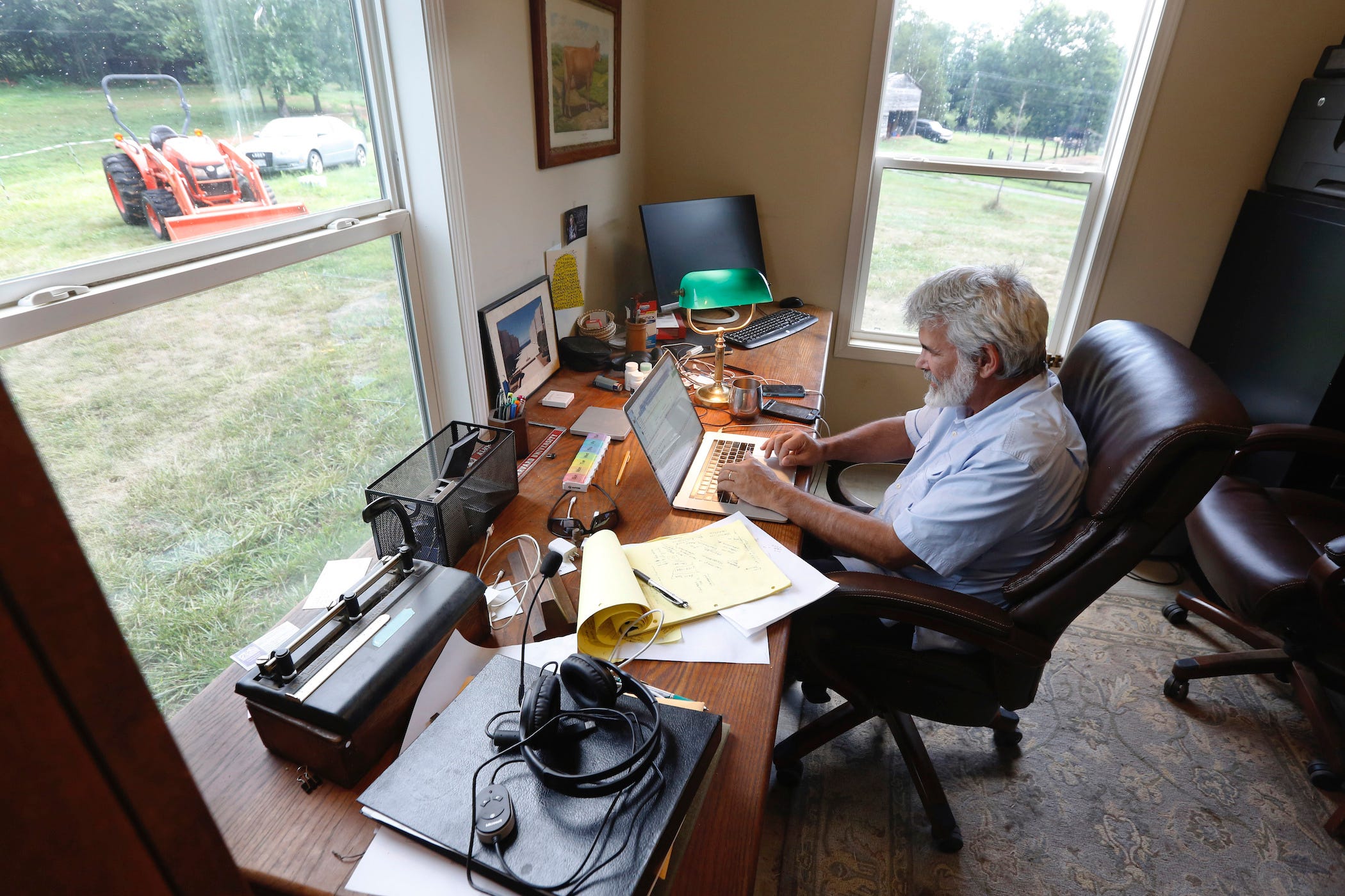 dr. robert malone working on his computer at his desk, farmland pictured out the window, with a tractor in view