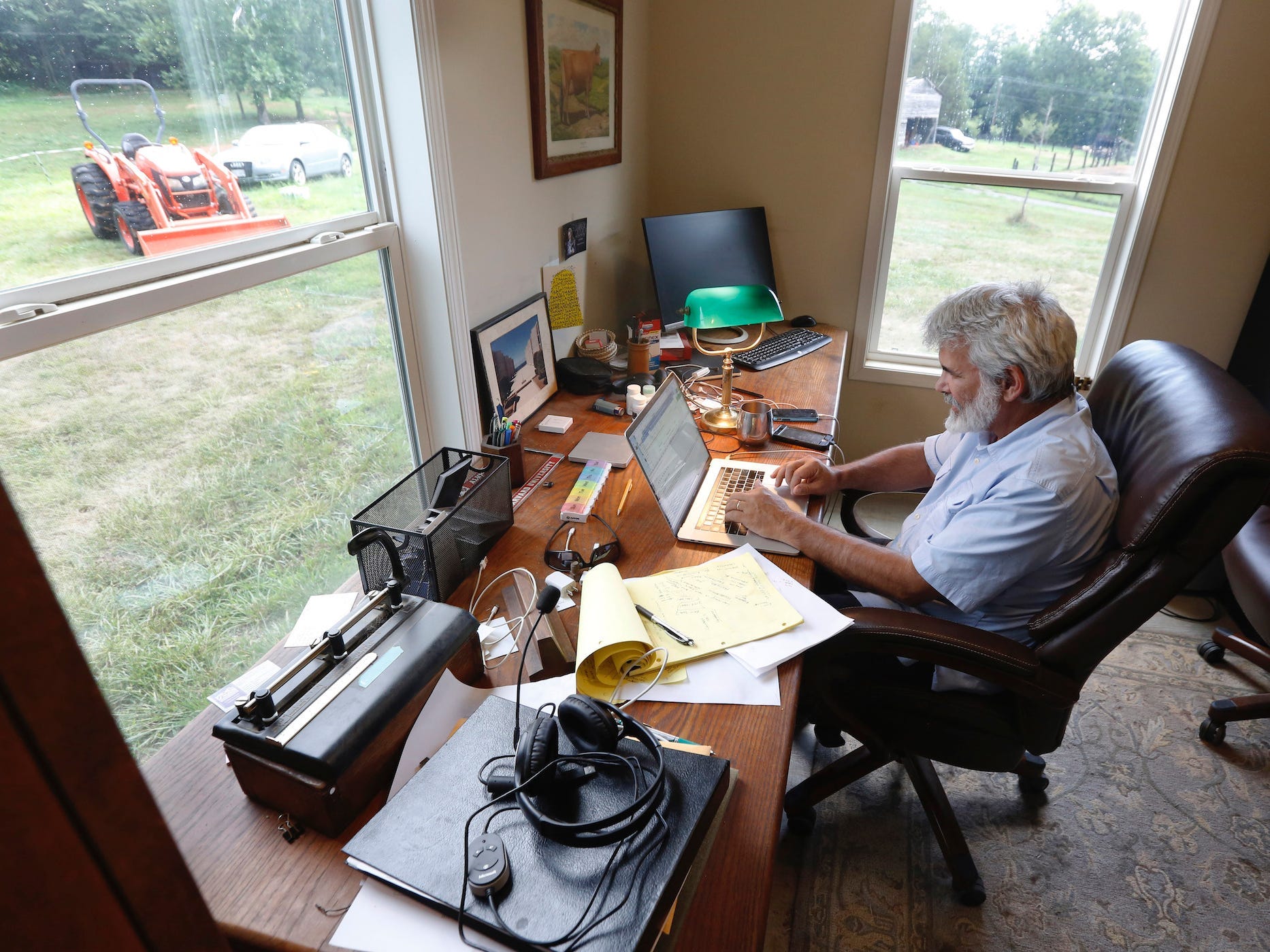 dr. robert malone working on his computer at his desk, farmland pictured out the window, with a tractor in view