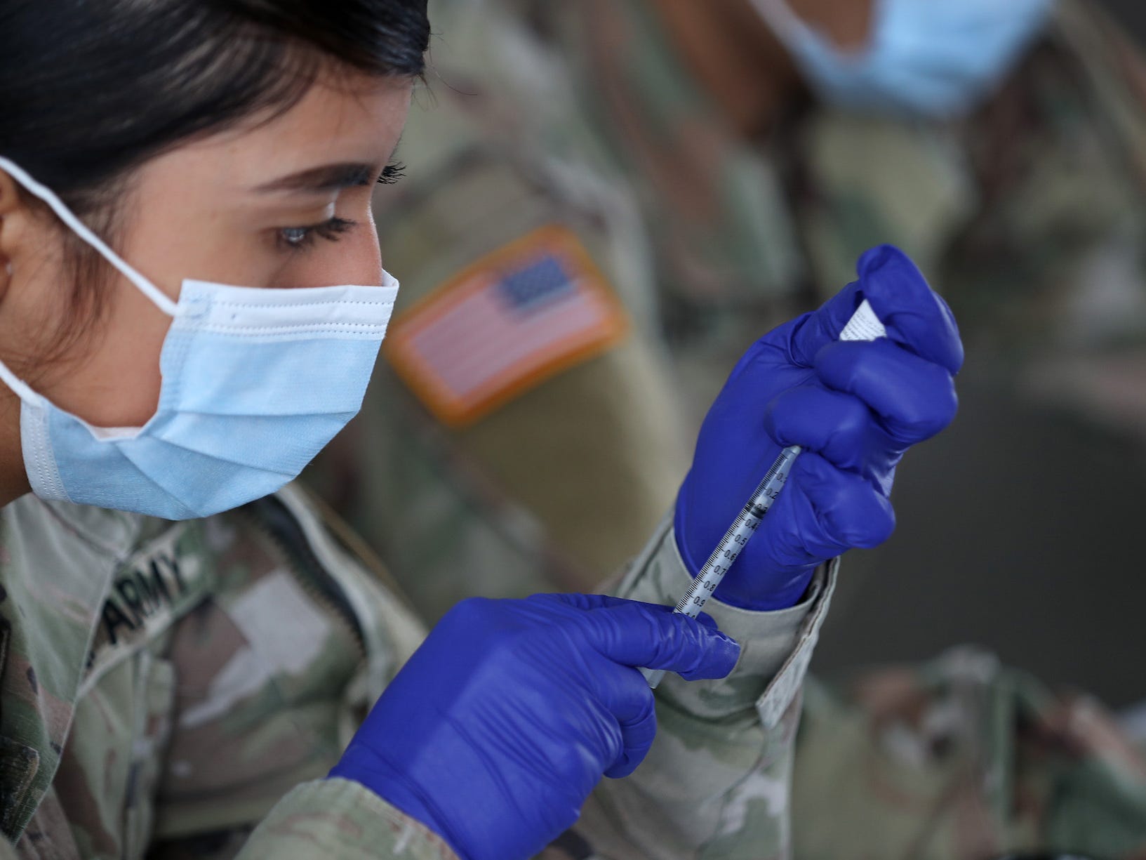 A woman wearing military garb a mask and gloves is measuring a dose of vaccine.