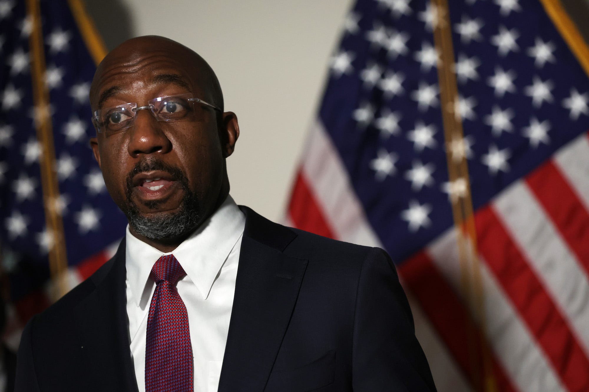 Suit-clad Democratic Sen. Raphael Warnock of Georgia speaks to reporters while standing against a backdrop of American flags at the US Capitol.