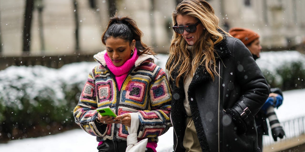 A guest (L) wears gold earrings, a neon pink oversized wool ribbed turtleneck pullover, a white / black / yellow / blue / pink square print braided pattern zipper high neck coat, a white leather clutch, black shiny leather large pants, a guest (R) wears a black leather with sheep interior zipper coat, khaki pants, a wool beige pullover, sunglasses, outside Ulla Johnson, during New York Fashion Week, on February 13, 2022 in New York City.