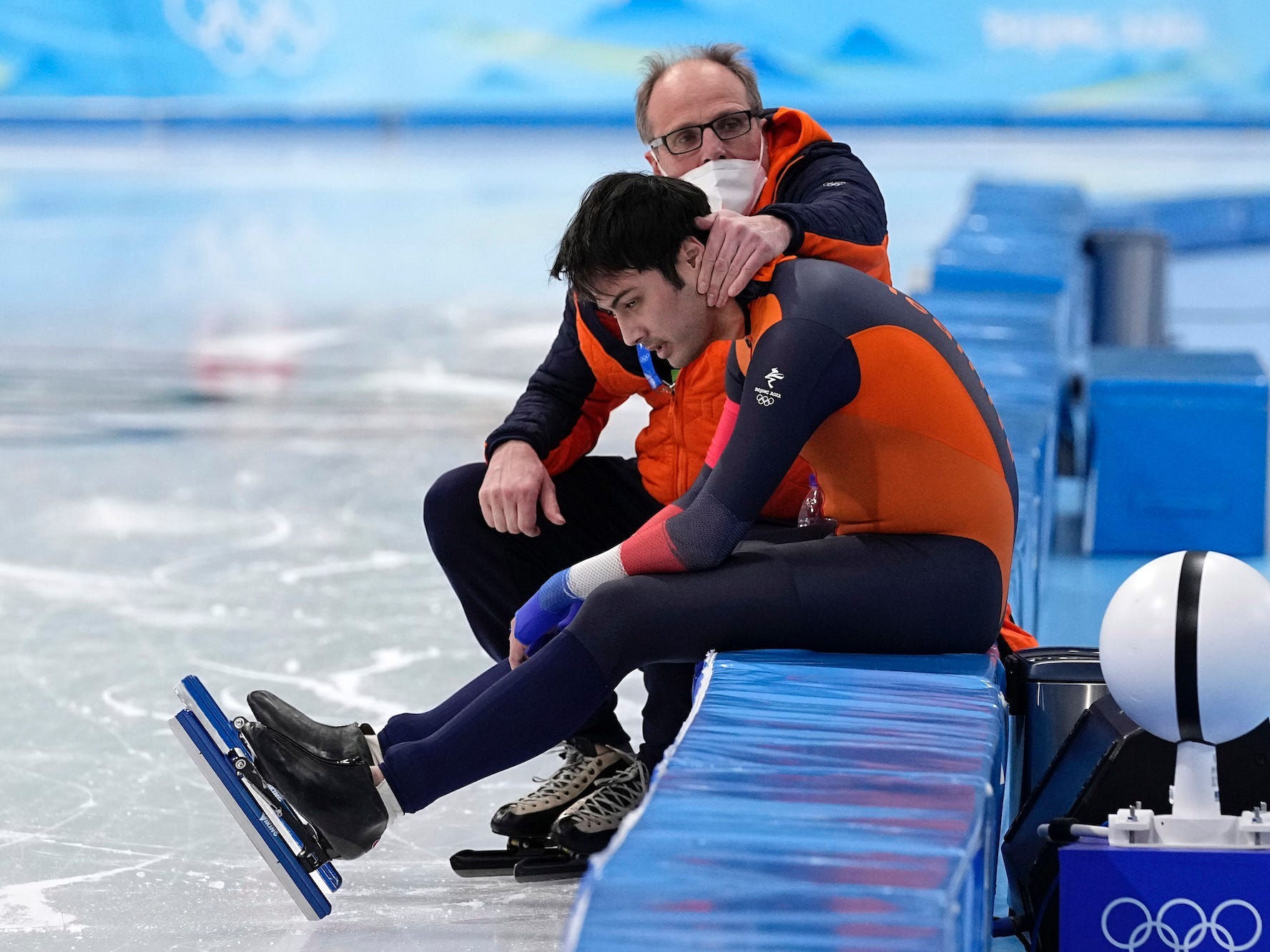 Kai Verbij sits on a bench on the ice as his coach puts his arm around him.