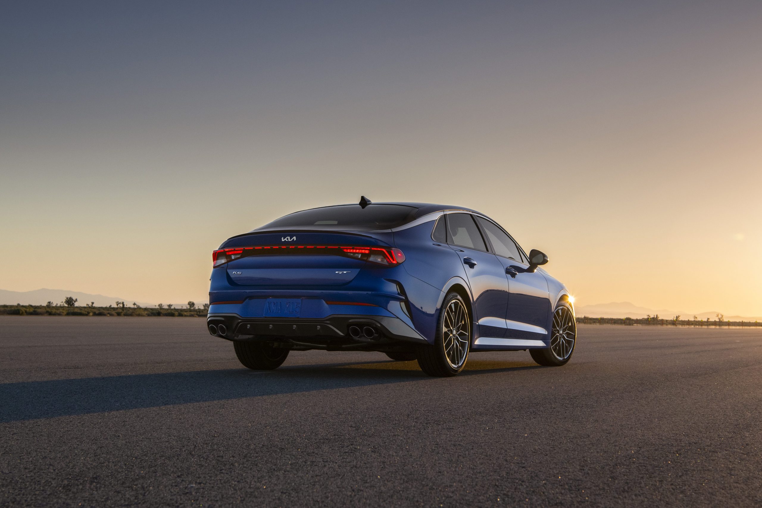 A rear view of a blue Kia K5 at sunset.