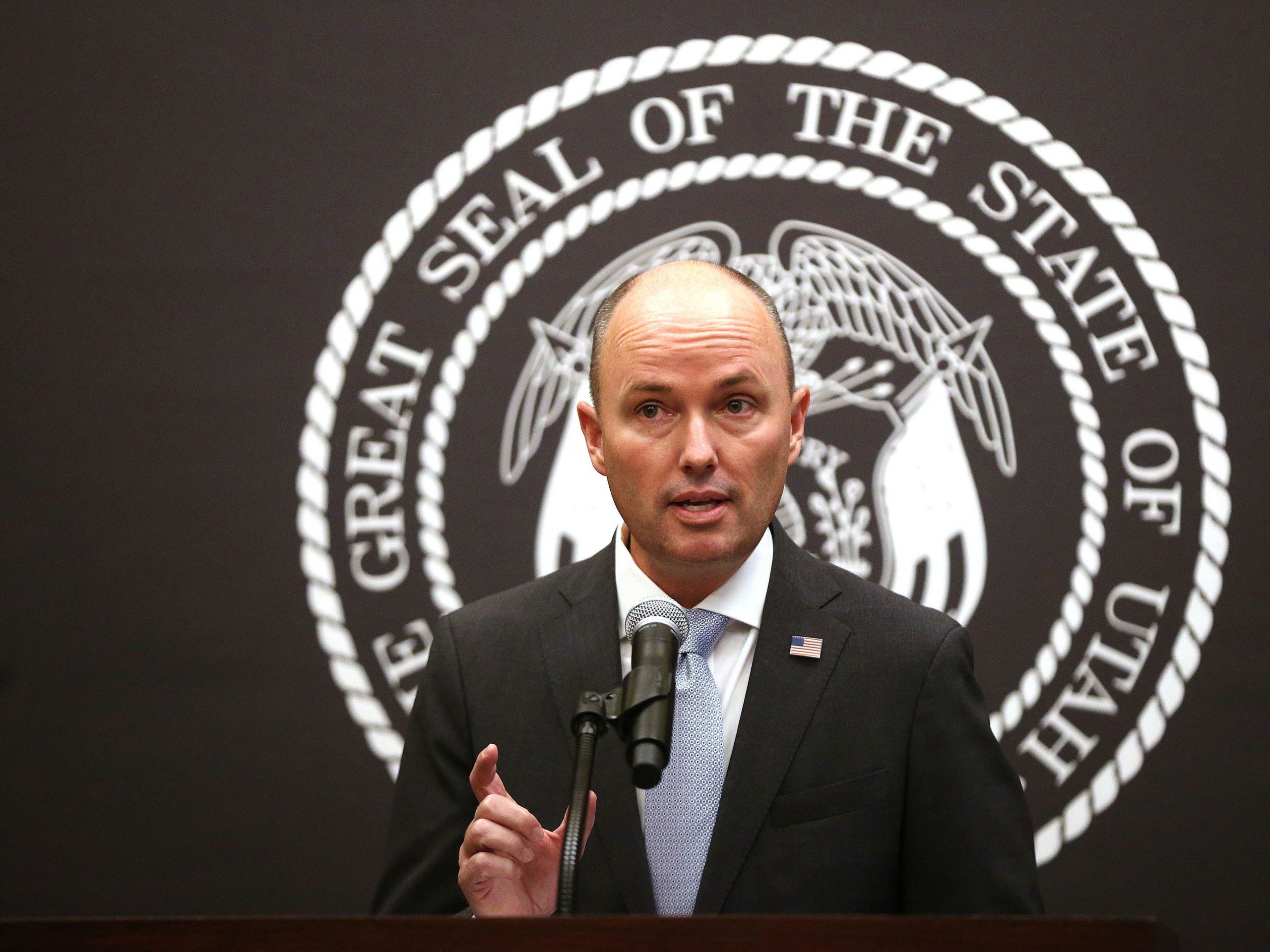 Spencer Cox in a black suit and blue tie speaking at a microphone.