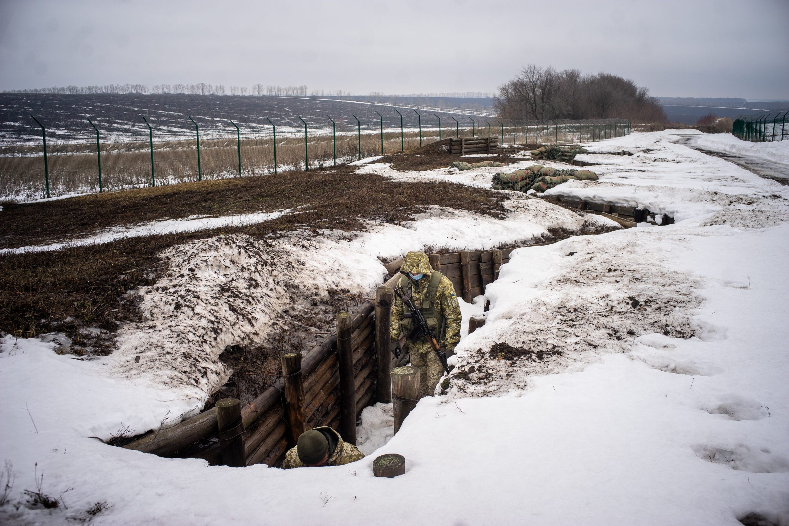 Trenches are seen in a desolate, snow-covered landscape near a long fence.