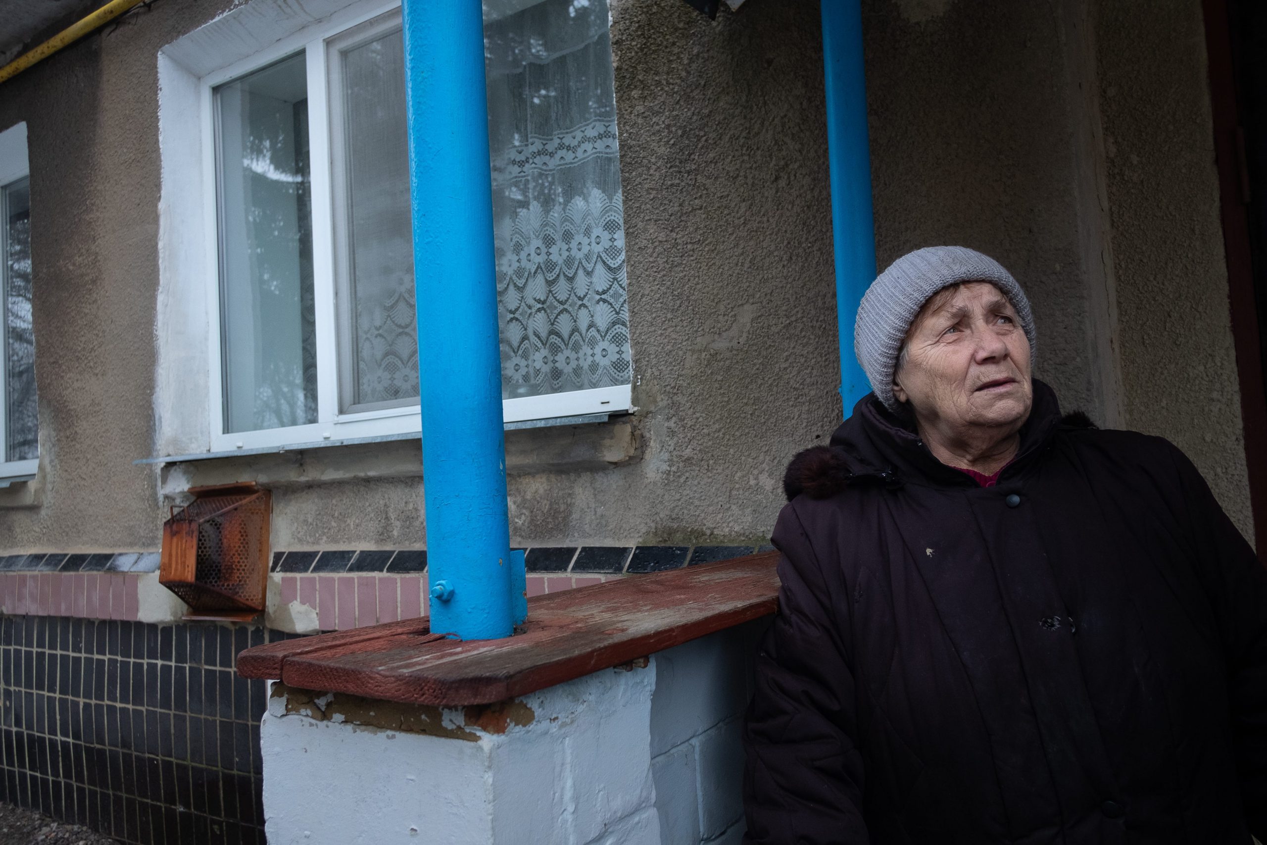 An elderly lady wearing a coat and hat stands outside a home.