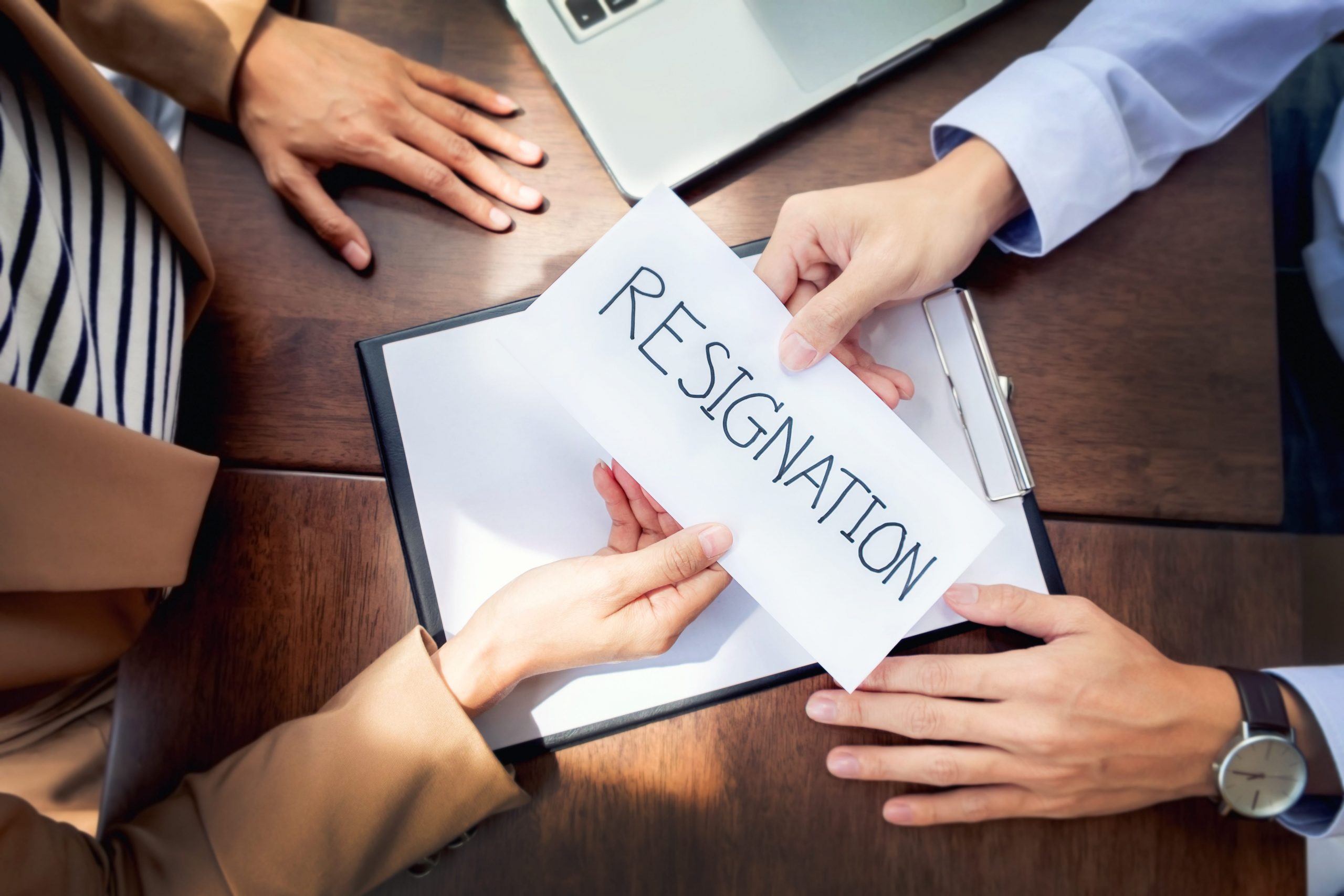 A top view of a desk with two people on either side of it, but only their hands are showing. One is handing the other a letter that says "resignation."