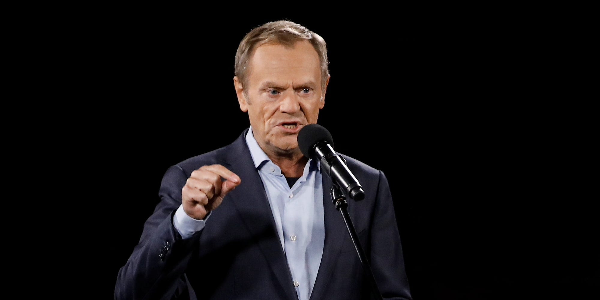 Donald Tusk looking angry speaking into a microphone against a black background.