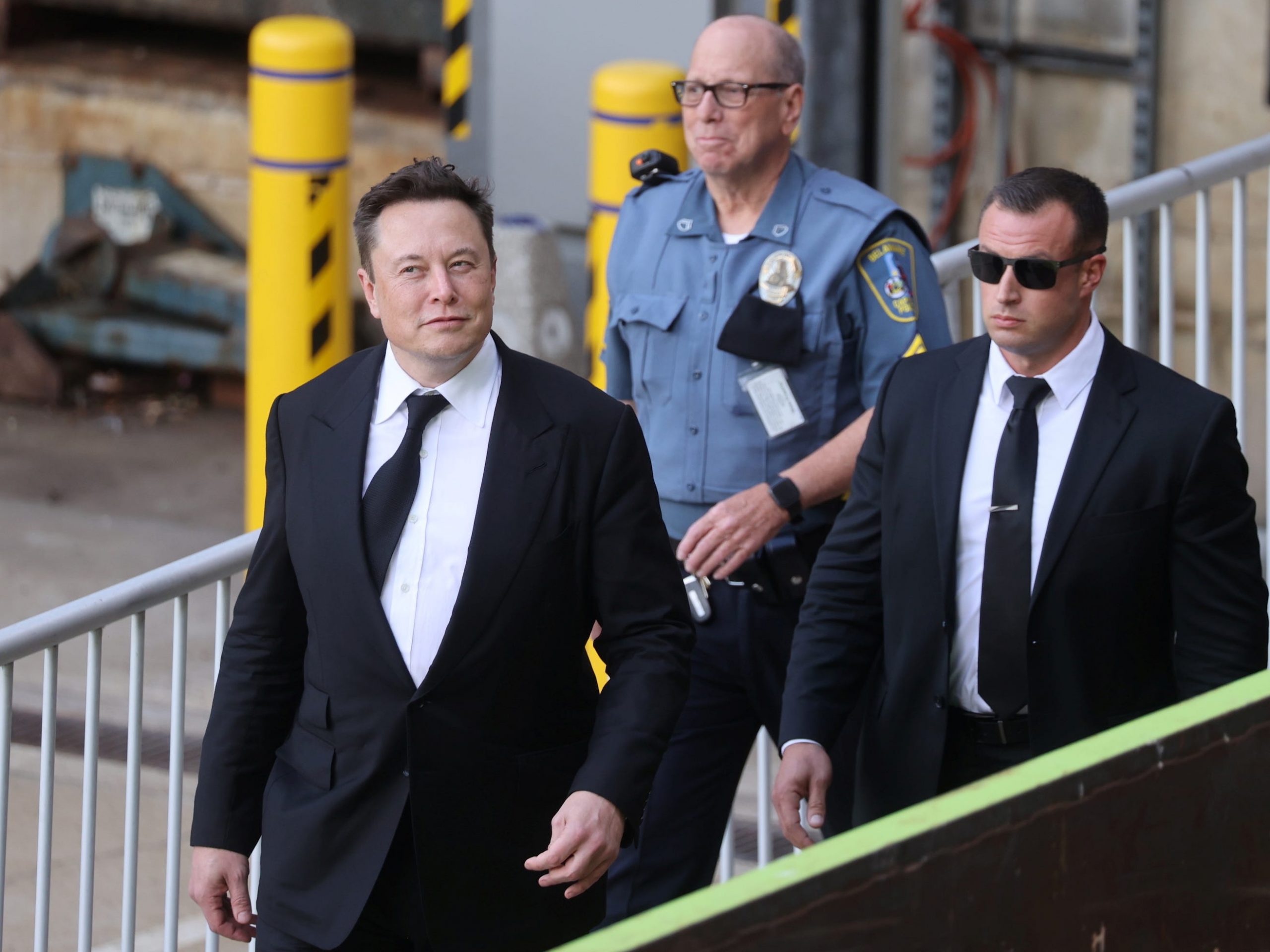 Elon Musk, wearing a black suit and flanked by another man in a black suit and a police officer, walking in front of a gray metal barricade.