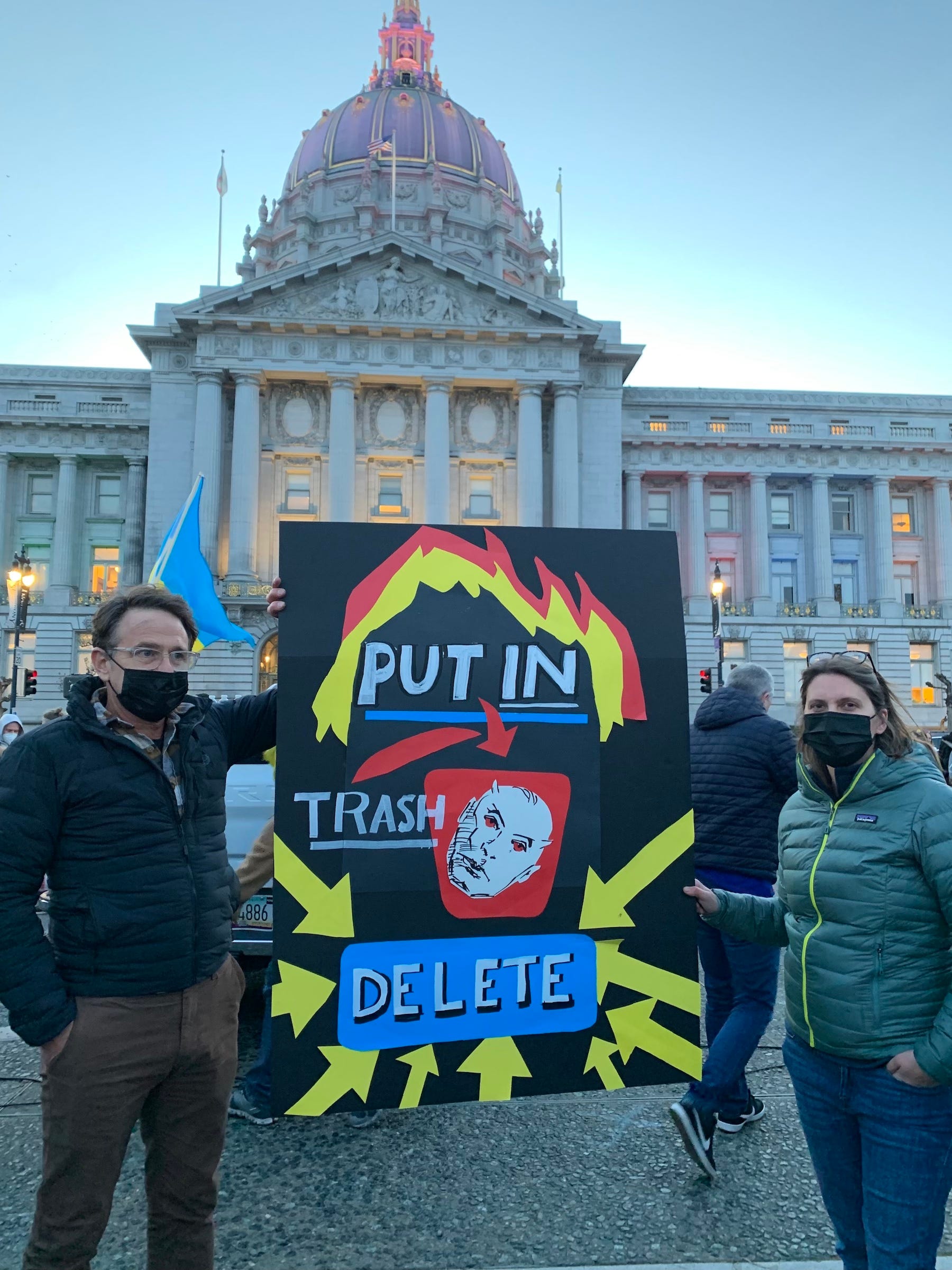 man and woman pose in front of san francisco city hall with sign that reads "put in trash delete" with putin's face