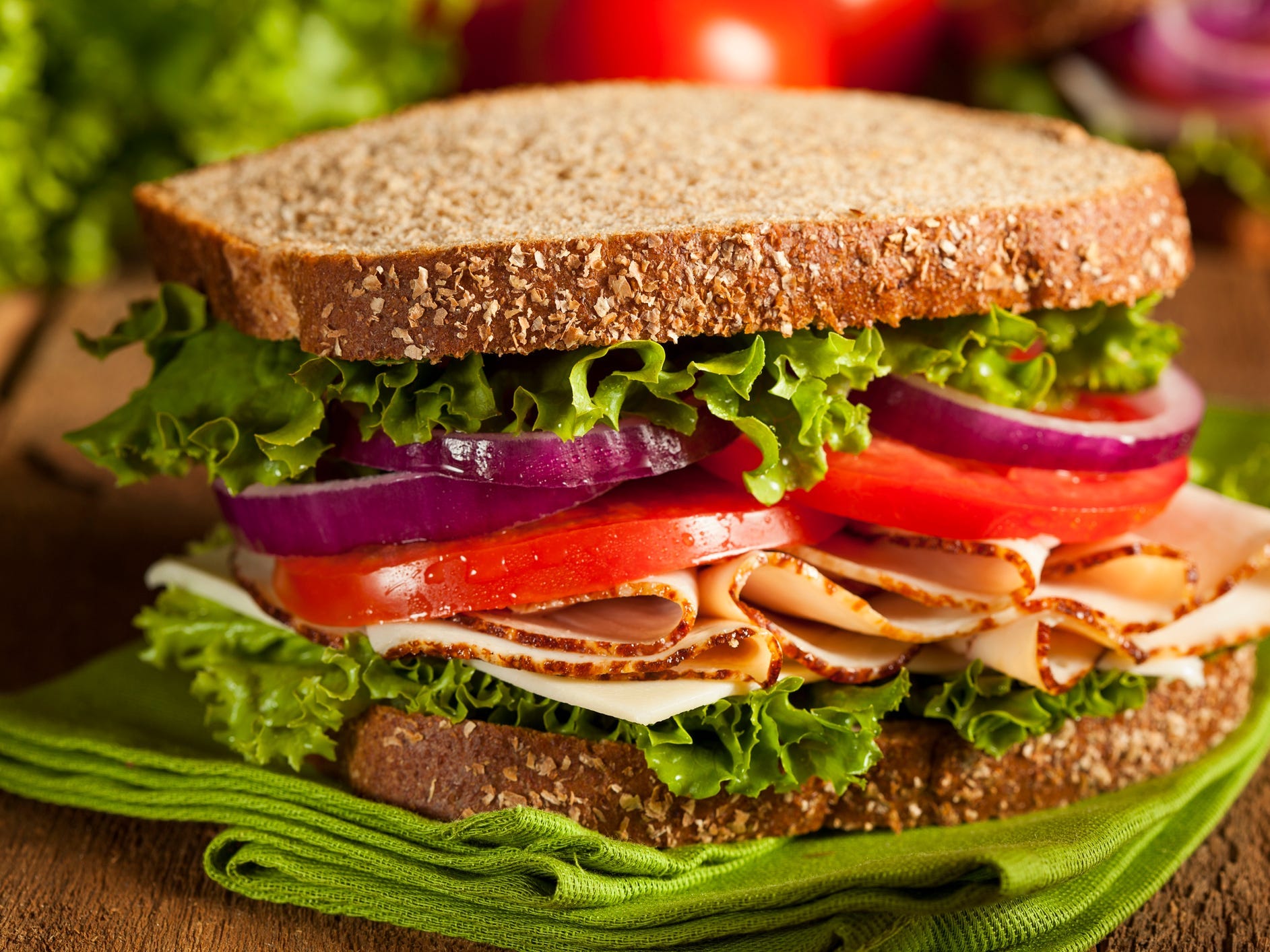 a turkey sandwich with whole grain bread, lettuce, tomatoes, and onions, resting on a green napkin atop a wooden table.