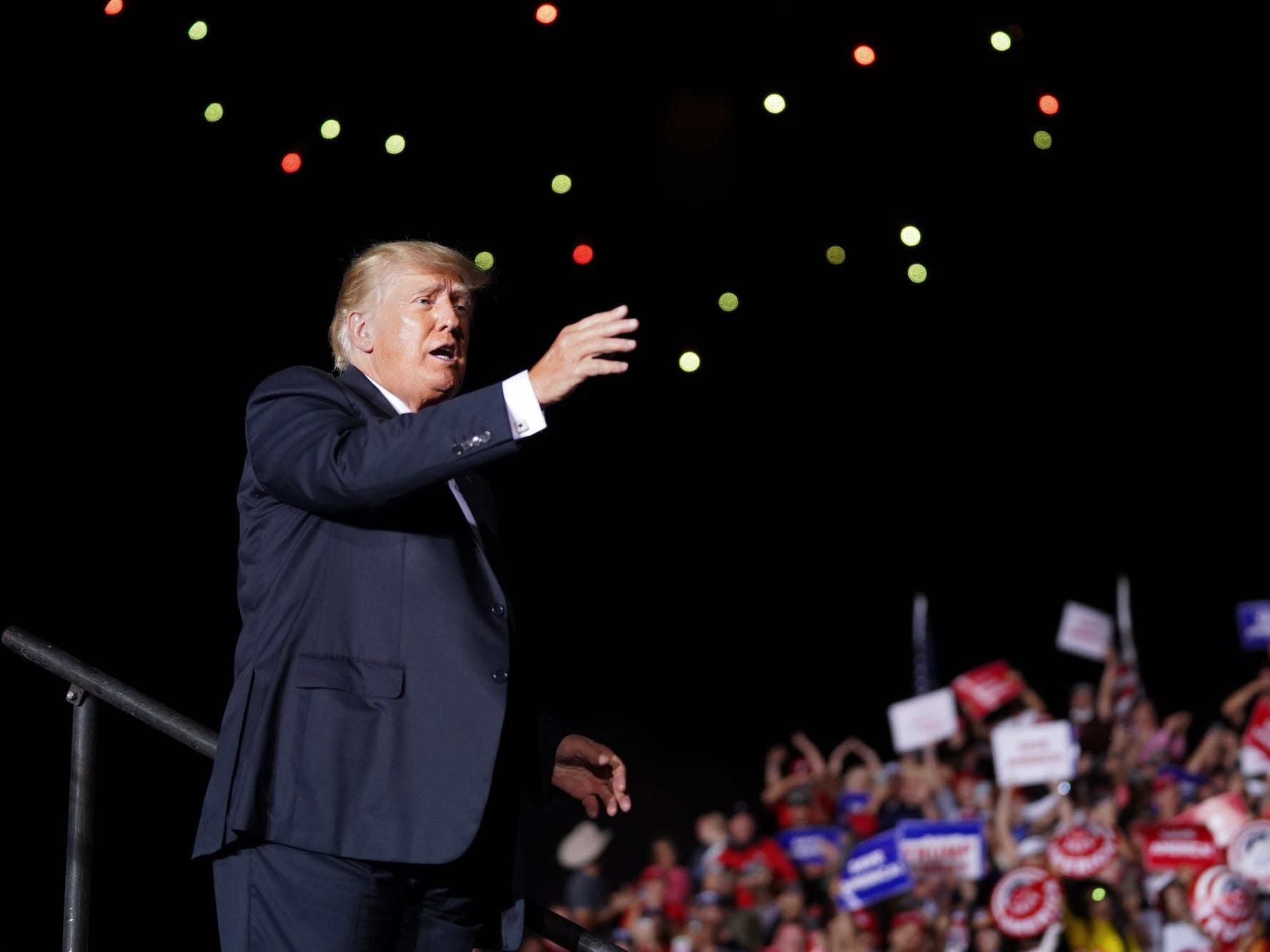 Former President Donald Trump, clad in a dark suit, his right arm extended upward as he stands against a starry sky, waves to attendees at a political rally in Georgia.