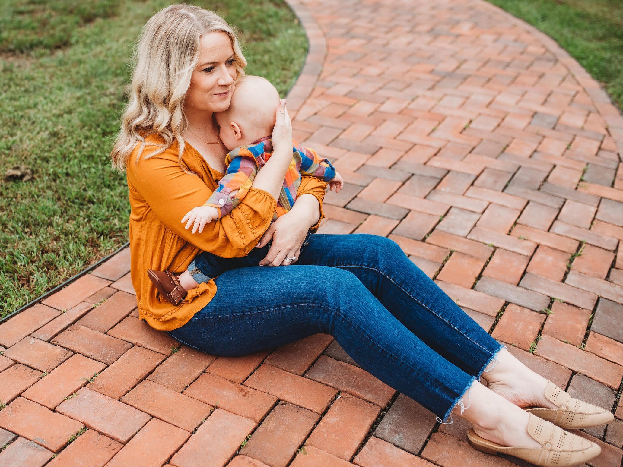 The author, a blonde woman, sits on a brick path outside. She wears jeans, an orange shirt, and holds an infant against her chest.