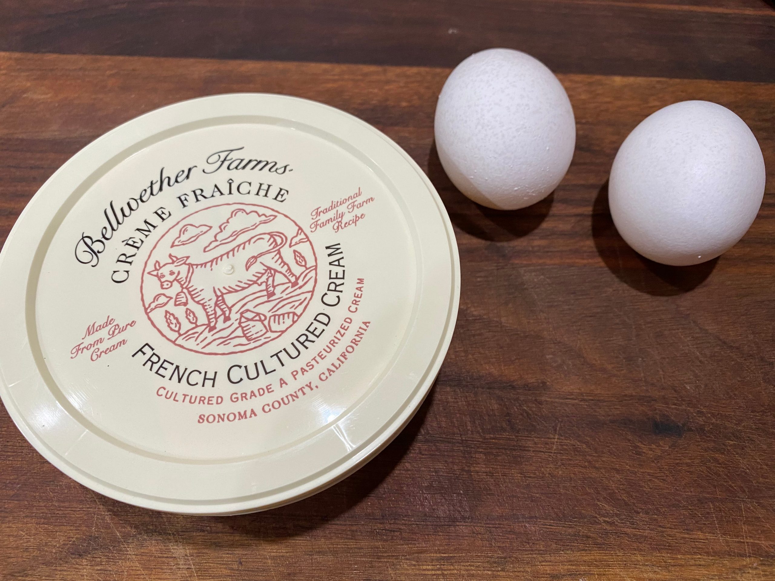 Can of crème fraîche next to two eggs with white eggshells on wooden cutting board