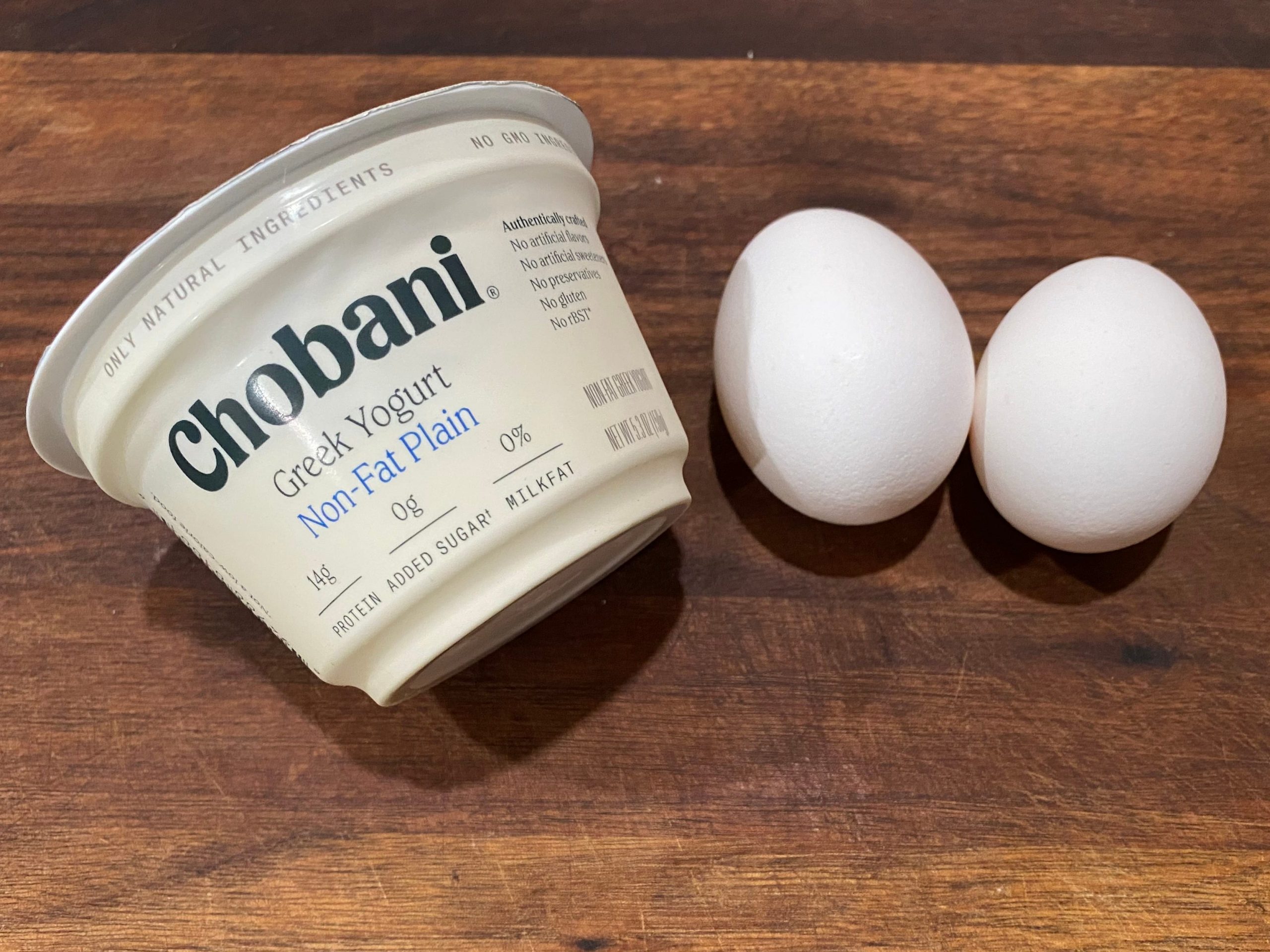 Cup of Chobani Greek yogurt and two eggs with white shells sit on a wooden cutting board