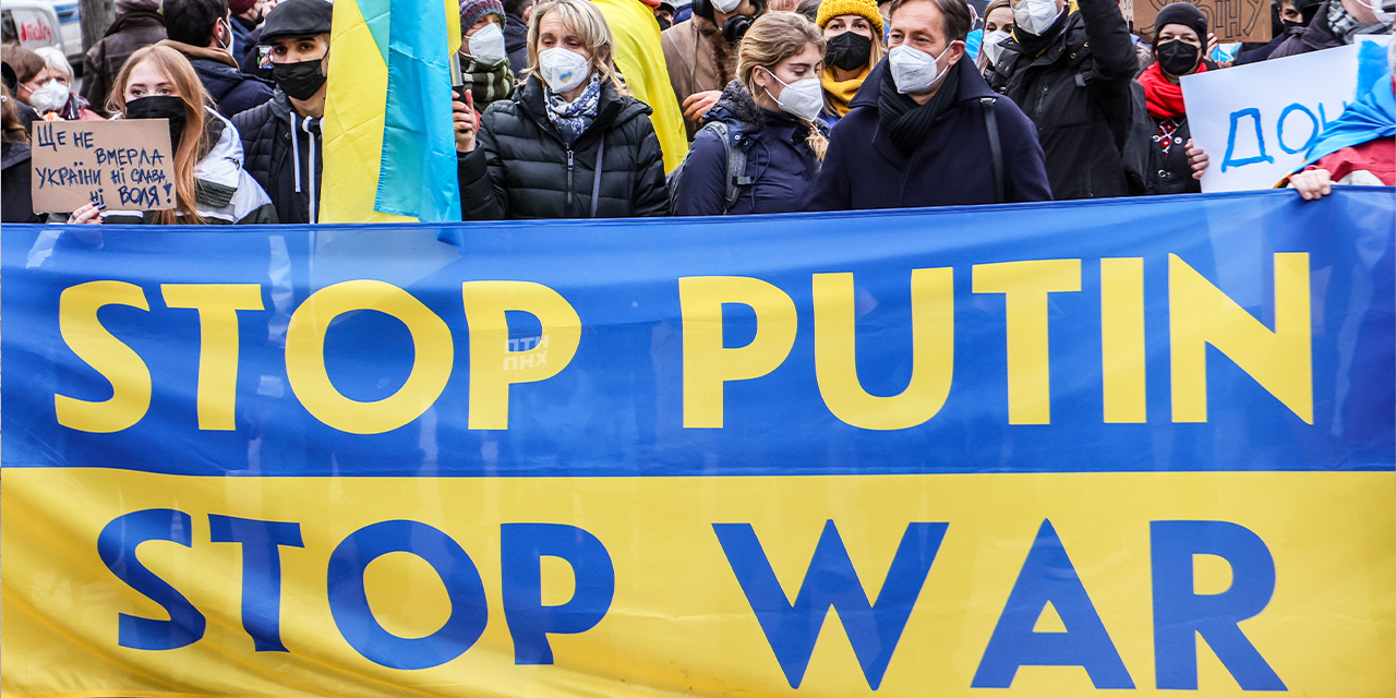 People hold a blue and yellow Ukrainian flag that says "Stop Putin Stop War" while wearing masks