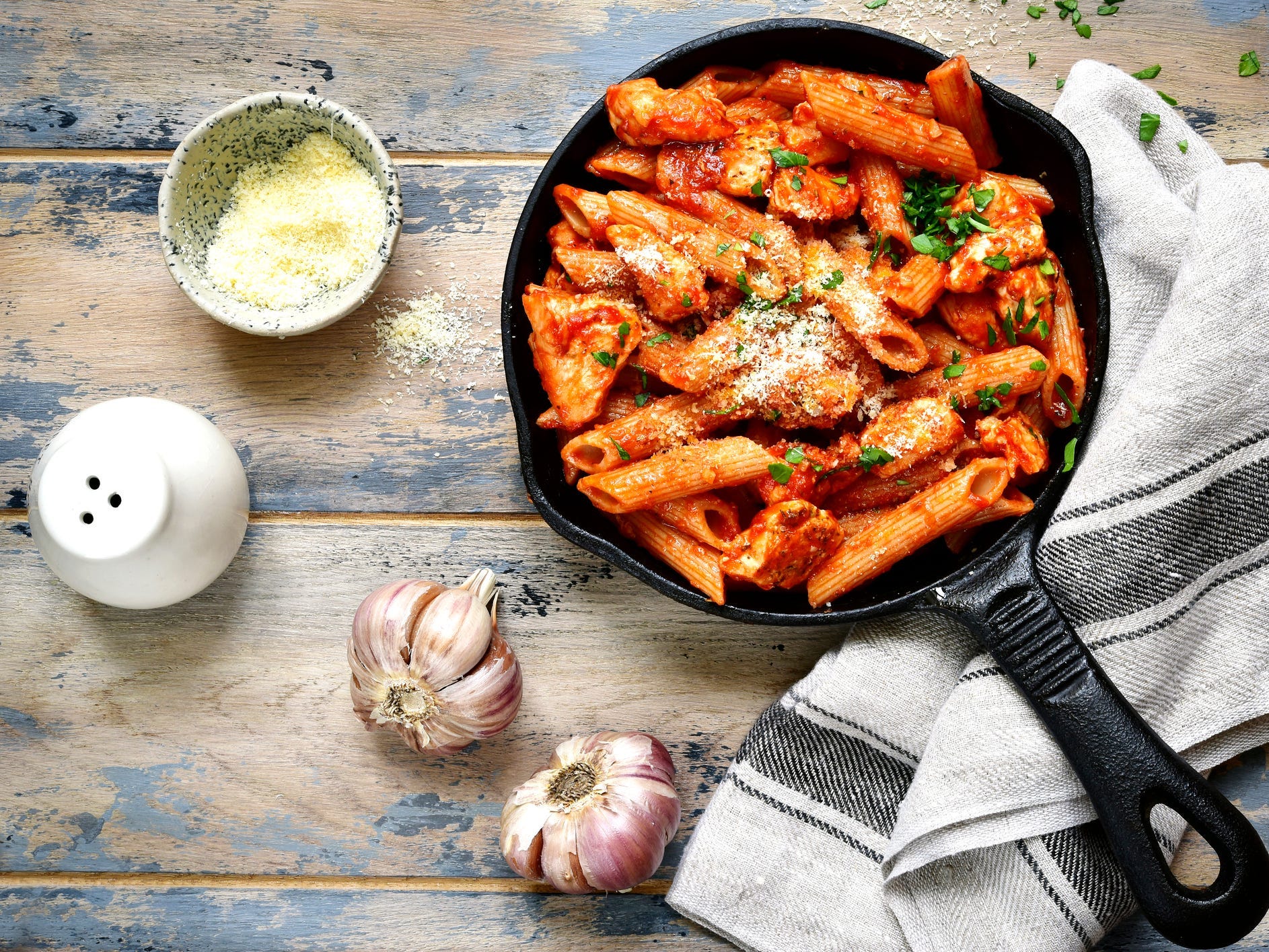 a cast iron skillet full of pasta with chicken, red sauce, herbs, veggies and cheese on a wooden table next to heads of garlic and seasonings