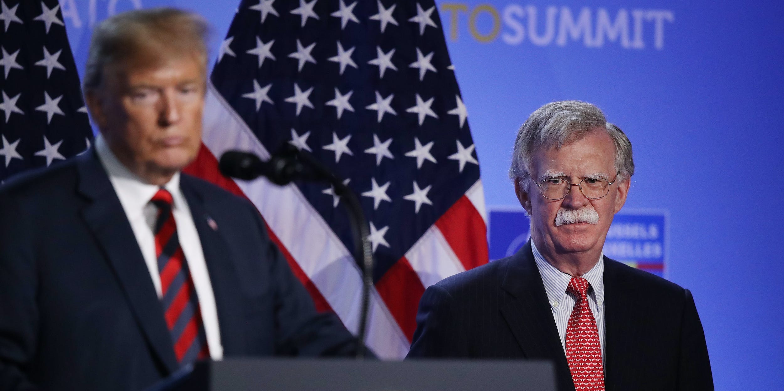 Donald Trump stands with his lips pursed as John Bolton looks directly into the camera from behind him.