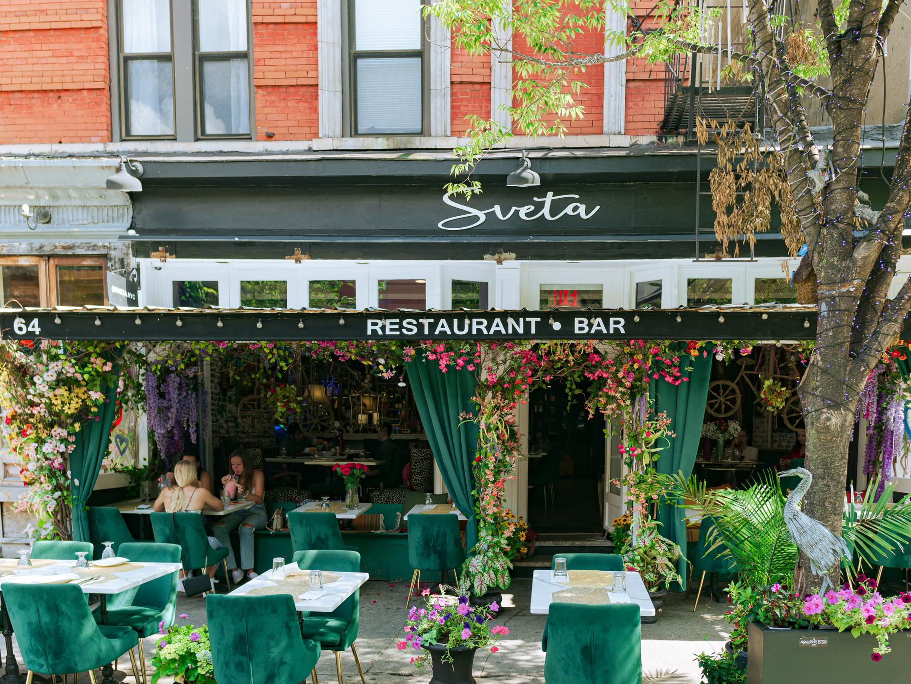 A photo of Sveta, a West Village restaurant that sells Ukrainian and Russian food. The building above the restaurant is red with two windows. The awning below that says "Sveta" in cursive script. Under that, it says "restaurant. bar." The chairs on the sidewalk in front of the building are green and the tables are white. There is a tree to the right of the photo.