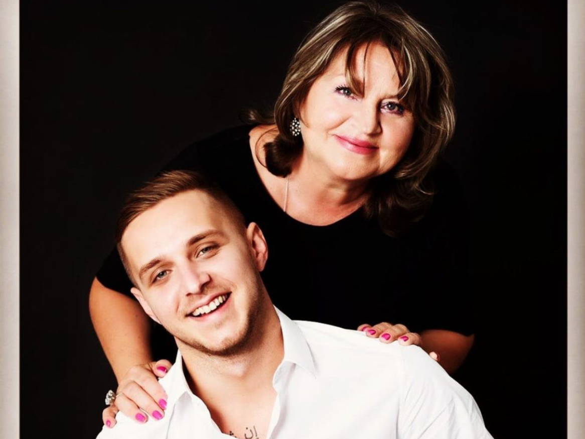 A mother and son. Sveta, the mom, has short brown hair and has her hands on her son, Alan's, shoulders. The background is black. He is wearing a white button-down.