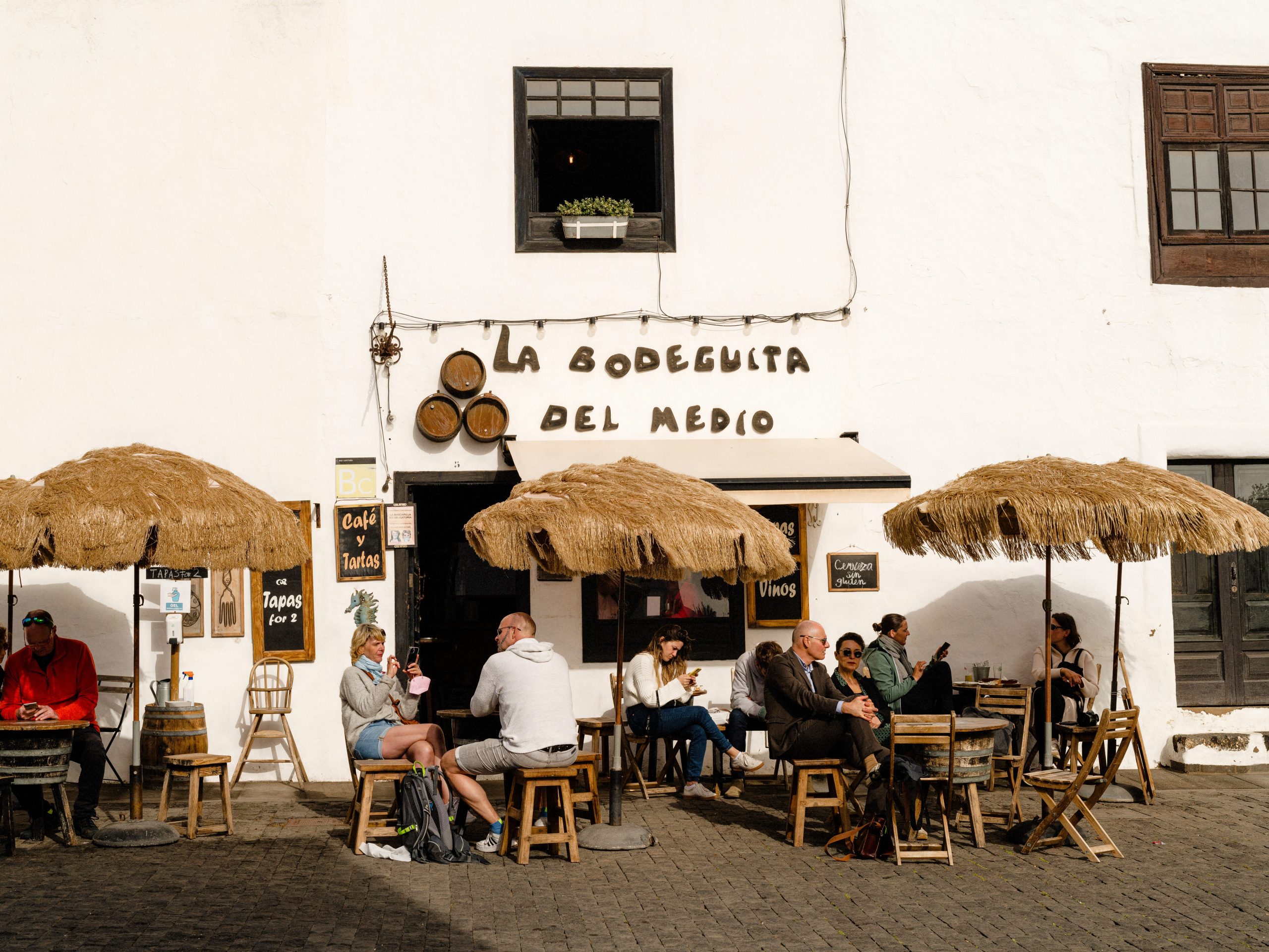 The outside of a restaurant in a white building with seating underneath grass umbrellas in front.