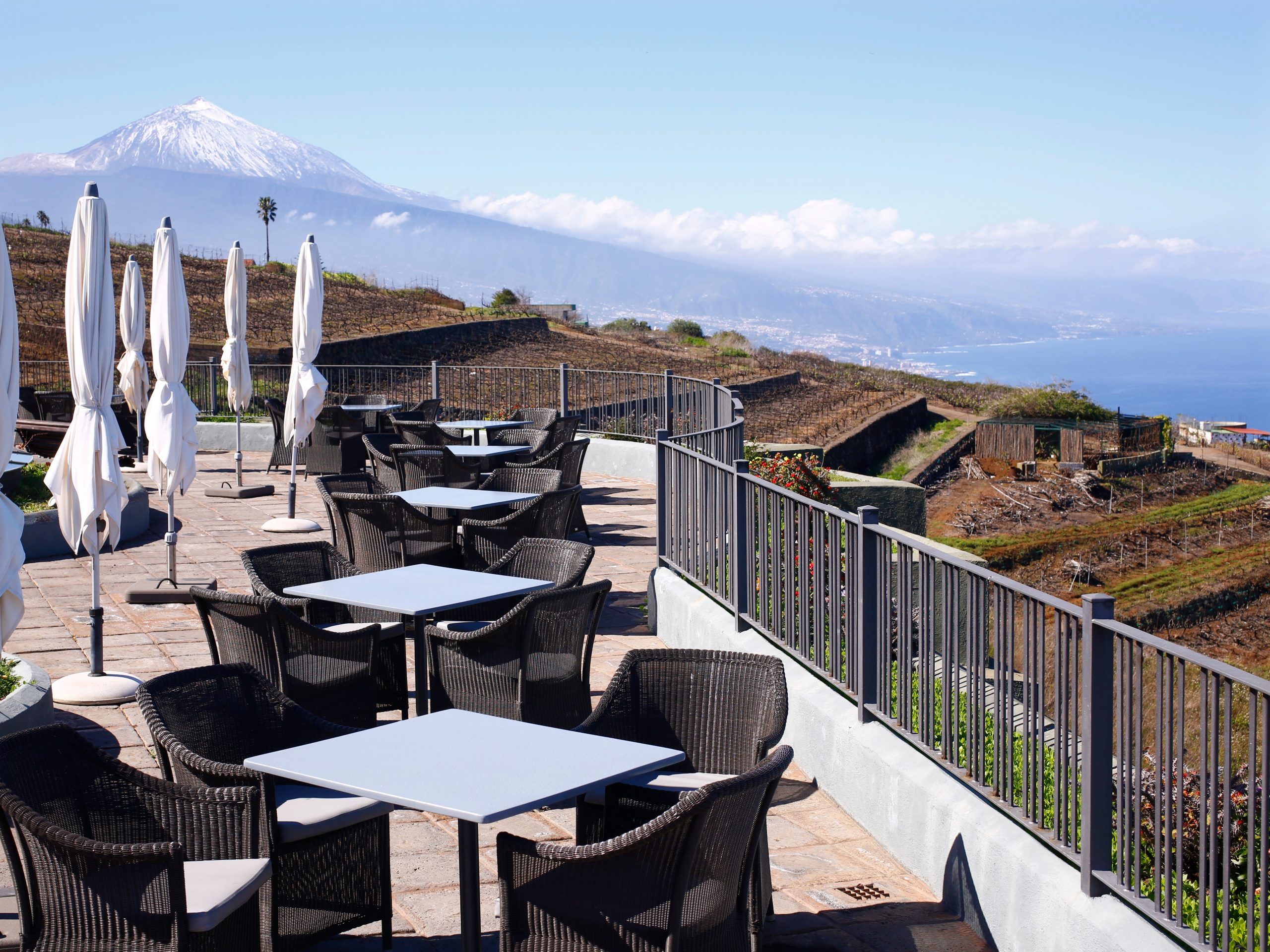 A balcony with several small tables surrounded by chairs next to closed white umbrellas.