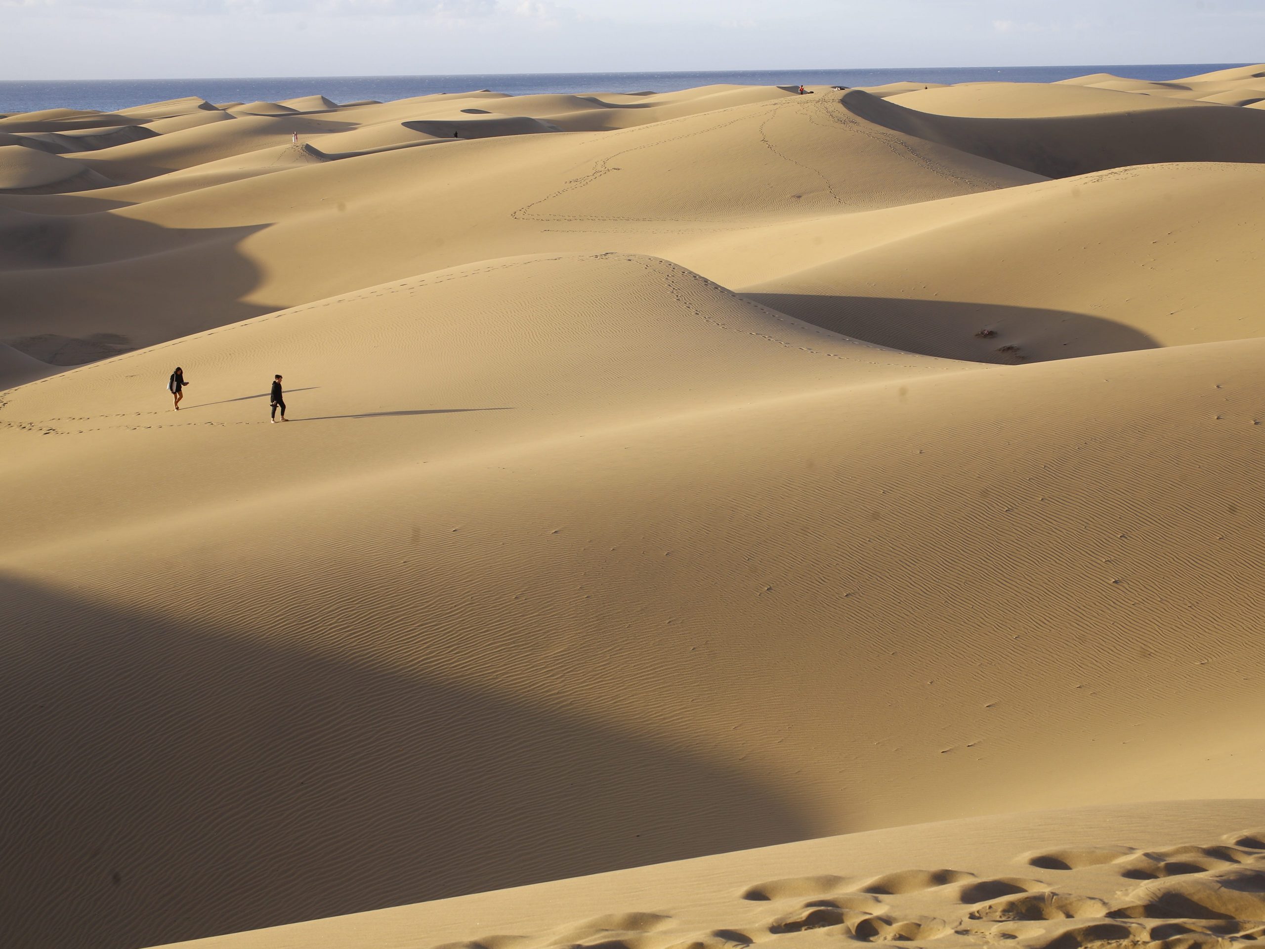 Two people walking across large sand dunes with the ocean in the far background.